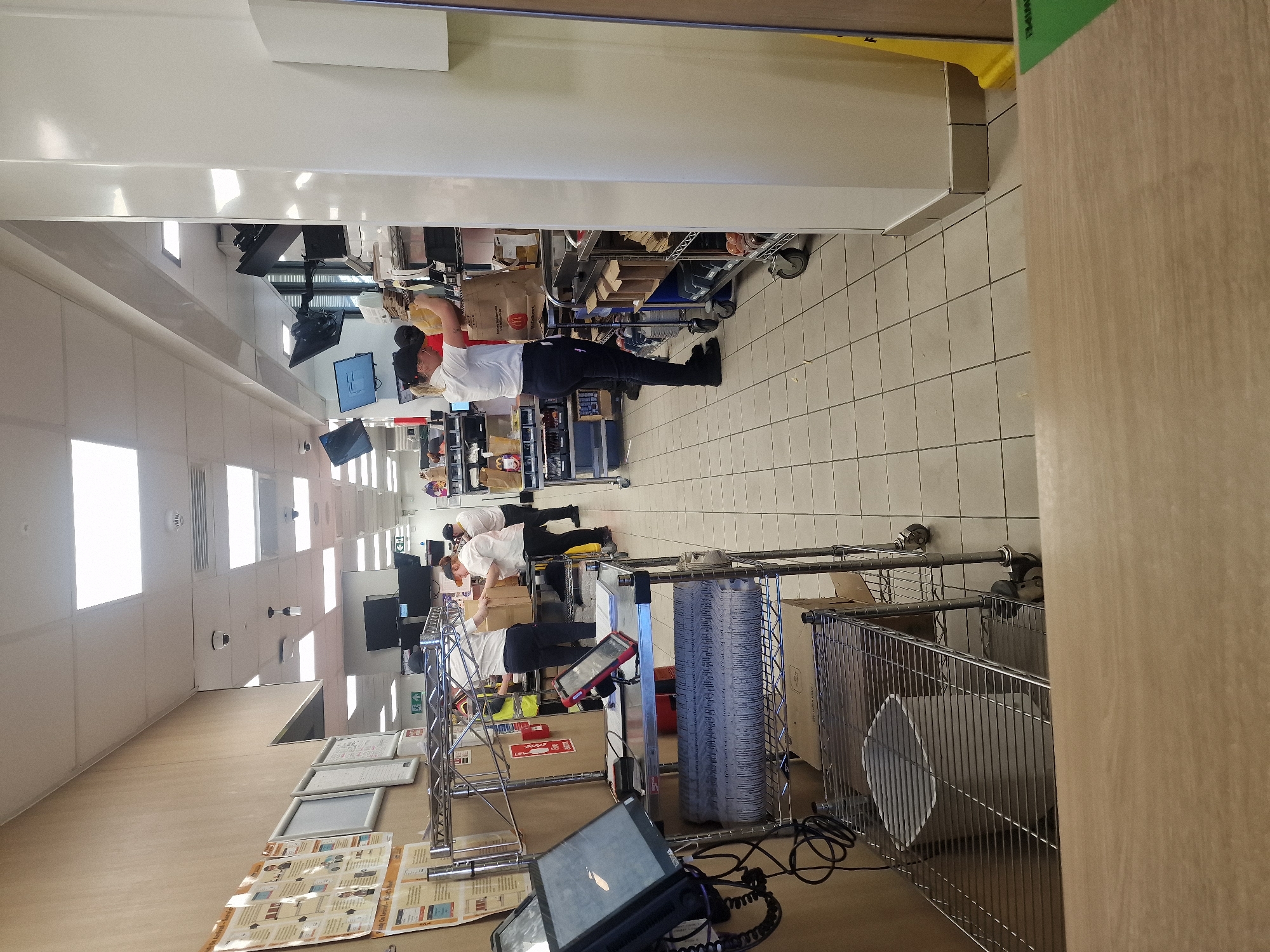 This image, rotated 90 degrees clockwise, captures an indoor back-of-house or operational area, likely for retail, logistics, or food service, given the mixed brand cues. The setting features a tiled floor, white walls, and bright artificial lighting from overhead fixtures.
At least three individuals, presumably employees, are visible. They are dressed in white short-sleeved shirts and dark trousers or jeans, suggesting a uniform. One person in the foreground (when rotated correctly) wears a dark cap and is holding a brown paper bag with the McDonald's logo on it, appearing to be engaged in packing or sorting. Another employee is leaning over a counter further back, also involved in work.
The area is equipped with various items:
* Metal shelving units and wire carts, some holding stacks of disposable cups/containers.
* Multiple computer monitors displaying information, indicating order processing or inventory management.
* Cardboard boxes, including a prominent red DHL Express box on the floor.
* A black tablet-like device in the immediate foreground, possibly for order management or scanning.
* A red handheld device, likely a scanner or payment terminal.
* Framed documents or posters are affixed to a wall or shelving structure.
The overall activity suggests order fulfillment, packing, or stock handling. The presence of both a McDonald's bag and a green label with "SNIPES" visible vertically on a wooden panel (which likely refers to a clothing/shoe retailer) indicates this could be a shared space, a dark store/kitchen, or a logistics hub serving multiple brands. The environment appears functional and geared towards efficient processing of goods. No specific time of day or emotional states are discernible beyond focused work.