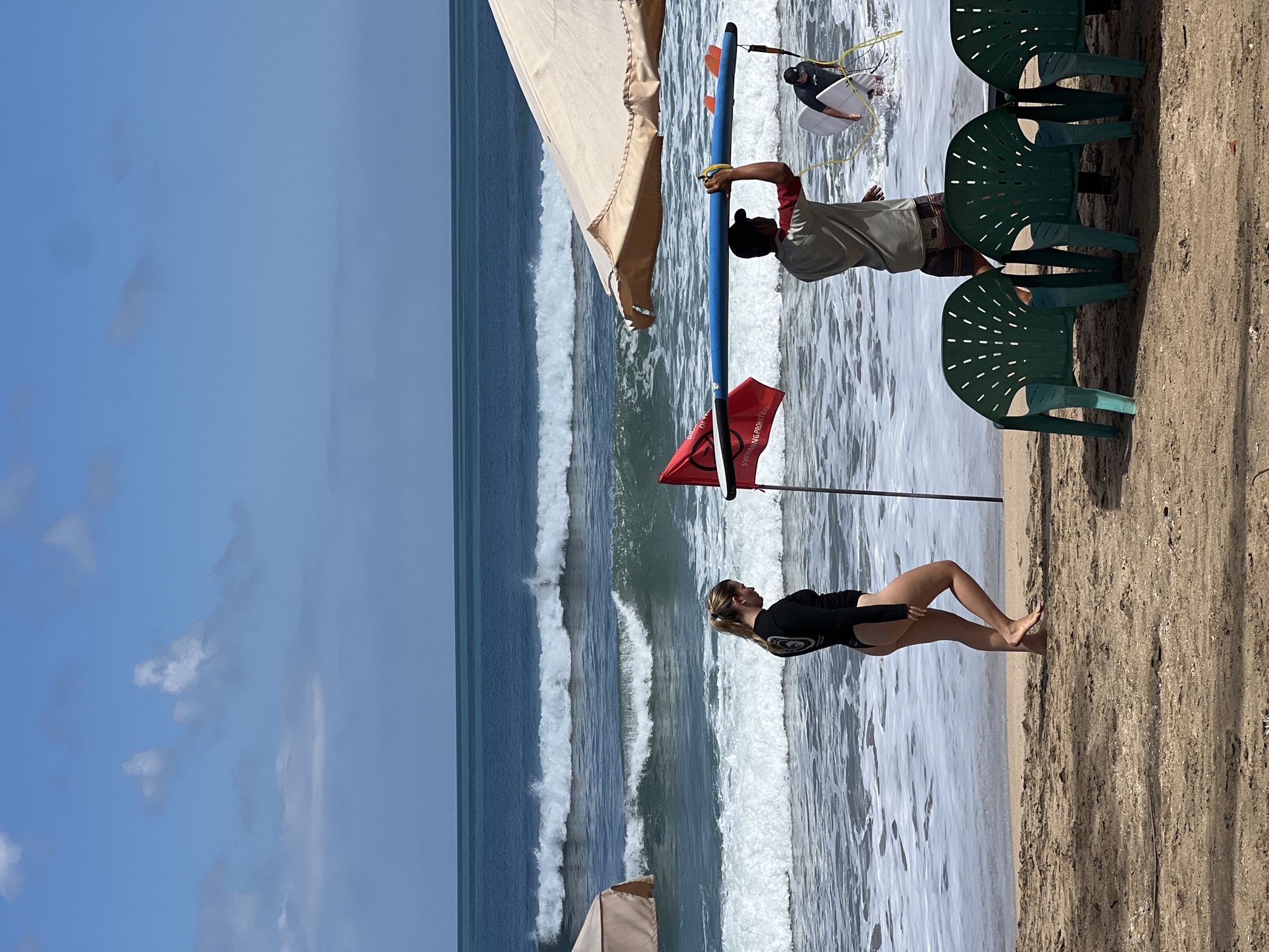 This image captures a beach scene in Kuta, Indonesia. In the foreground, three green plastic chairs are lined up on the sandy beach. To the right of the chairs, a man is carrying a blue surfboard overhead. He is wearing a red and beige shirt with dark patterned shorts. Behind him, another man is entering the water with a white surfboard. In the middle ground, a woman in a black swimsuit is walking along the shore, facing left. A red flag, with the words "SWIMMING PROHIBITED" printed on it, is planted in the sand near the man with the blue surfboard. The ocean in the background features moderate waves with white foam. The sky above is blue with scattered white clouds. Two large, light-colored umbrellas are visible on the beach, one to the left and one to the right, providing shade.