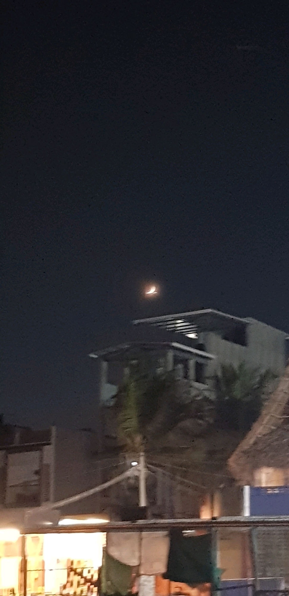 A nighttime photograph captures a crescent moon illuminated in the dark sky above a multi-story building. The building appears to have a modern architectural design with horizontal louvers on its upper level. Palm trees are visible near the building, suggesting a tropical or subtropical environment. In the foreground, a covered structure with hanging fabrics and bright lights indicates a marketplace or an outdoor dining area. The overall scene depicts a dimly lit urban or semi-urban environment at night in Thandiriyan Kuppam, India.