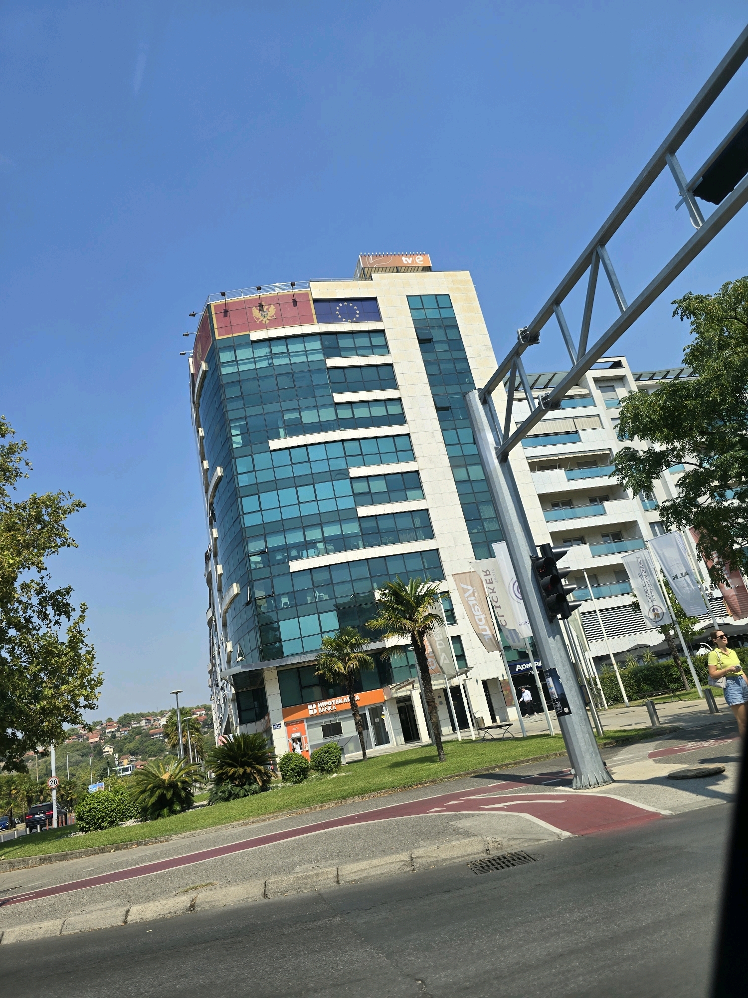 An eye-level, wide shot captures the facade of a modern, glass-fronted office building on a sunny day in Podgorica, Montenegro. The building features a prominent Montenegrin flag and a European Union flag at its apex, suggesting official or diplomatic significance. The lower levels of the building house various businesses, including "Hipotekarna Banka" and "Admiral," with flags displaying their logos visible on the sidewalk.
In the foreground, a traffic light pole stands to the right of the frame, with a traffic signal displaying red and yellow lights. The road in front of the building is marked with red directional arrows and white lines, indicating a junction. Several palm trees and lush green bushes line the walkway, adding a touch of tropical flora to the urban landscape.
To the right of the frame, a woman in a yellow top and denim shorts stands near the building entrance, looking towards the street. In the background, residential buildings are visible on a hill overlooking the city, further emphasizing the urban setting. The sky is a clear, vibrant blue, indicating good weather conditions. The overall impression is one of a bustling, modern city, with elements of both corporate and residential life coexisting.