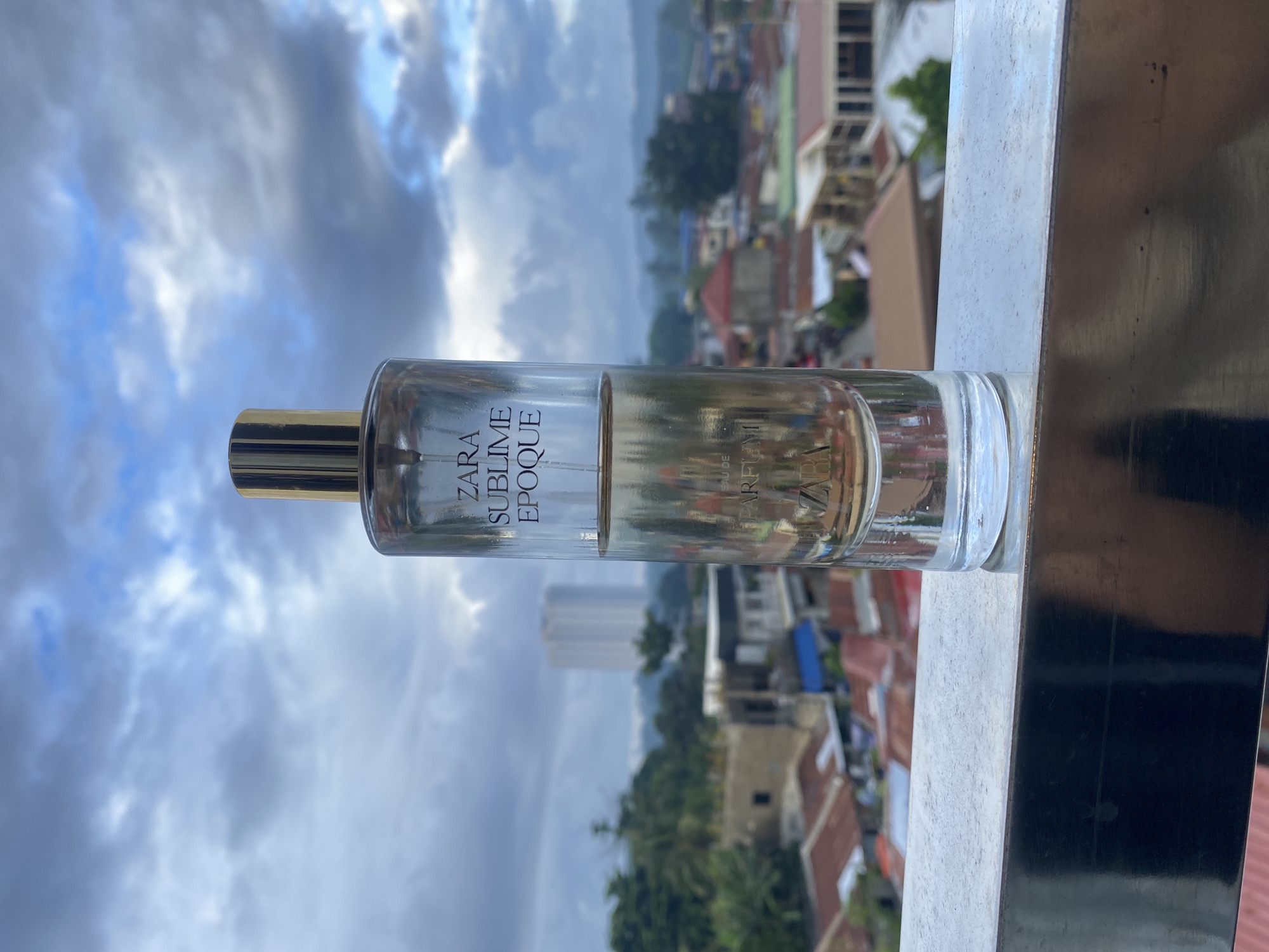 A clear glass perfume bottle with a gold cap is centered in the foreground, resting on a metallic railing. The bottle is labeled "ZARA SUBLIME EPOQUE" and "EAU DE PARFUM". The background depicts a cityscape under a cloudy sky, featuring houses with reddish-brown roofs, trees, and a tall building in the distance. The location is identified as Davao City, Philippines.