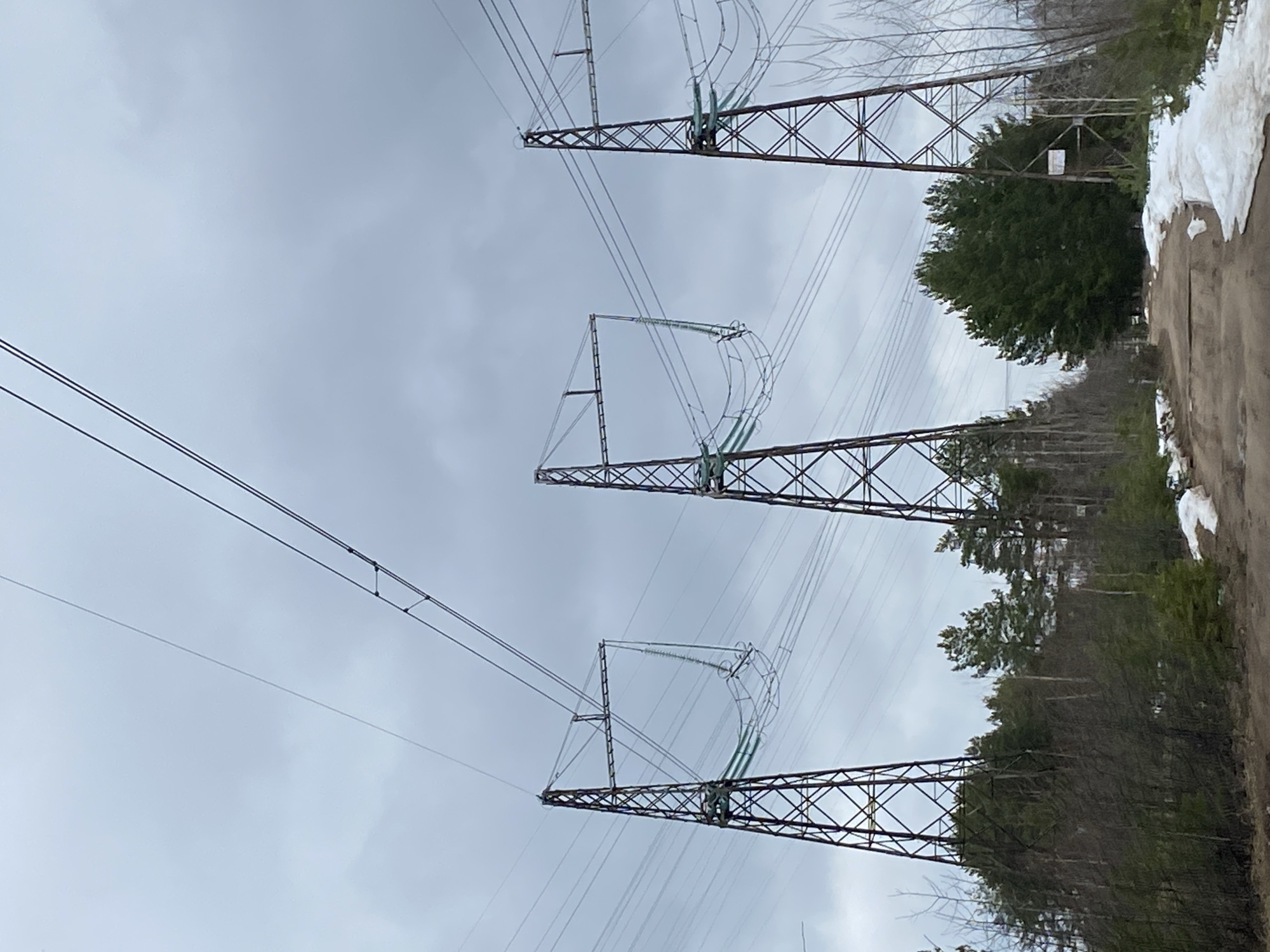 The image shows three power line towers under a cloudy sky. The towers are made of metal and have a lattice structure. Power lines are strung between the towers. The towers are situated next to a dirt road with some snow on the side. Trees are visible behind the towers. There is no evidence indicating this location is in Noviy, Russia.
