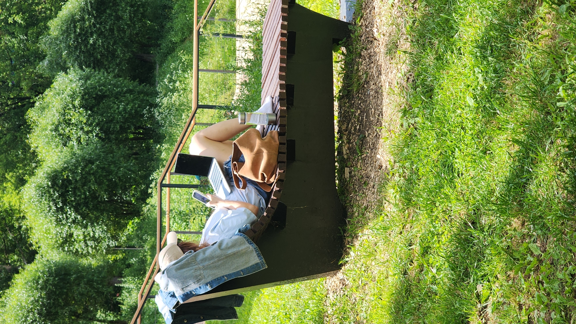 A person sits on a slatted wooden bench in a park, holding a smartphone and a laptop. The person is wearing a light blue shirt, denim shorts, and white socks. A brown tote bag and a clear water bottle are placed on the bench next to them. A denim jacket is draped over the side of the bench. The setting is outdoors with lush green trees and grass, suggesting a sunny day. The person appears relaxed and engaged with their devices, possibly working or studying remotely. The background shows dense foliage, indicative of a natural, possibly wooded area. There is no visible text in the image. The overall impression is one of a peaceful, productive moment spent outdoors in a park.