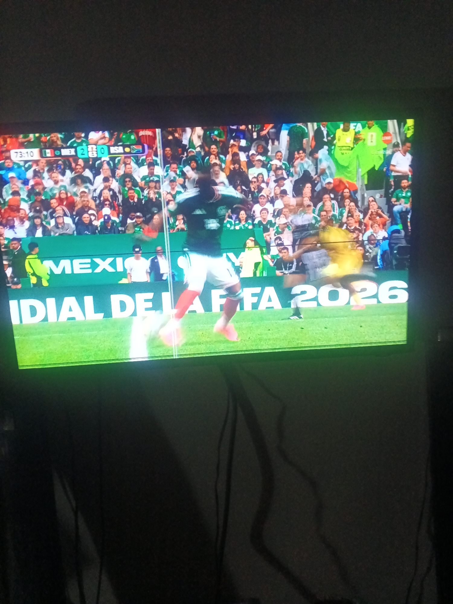 The image shows a wide-angle view of a soccer game on a television screen. The score is displayed as 2-0, with Mexico leading. The game is in its 73rd minute. The words "MEXICO" and "IDIA" are visible on a green backdrop behind the players, along with the text "FA 2026". A soccer player in a dark green uniform is in motion on the field, with another player in yellow blurred in the background. The audience is visible in the stands, wearing various colors, many of them in green. The television screen is reflecting some ambient light, and the overall image quality is slightly grainy.
