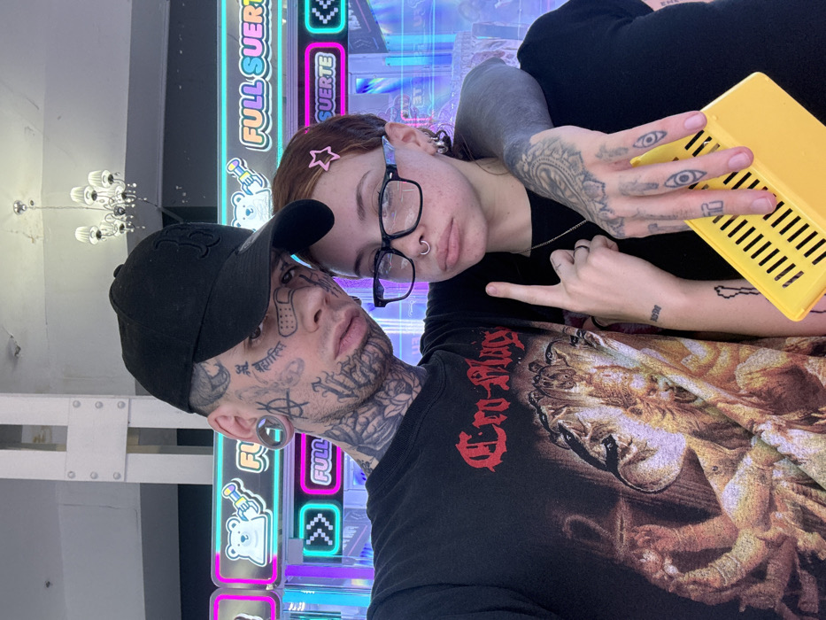 A tattooed couple poses closely. The man wears a black baseball cap and a black shirt featuring art. His face is heavily tattooed. The woman wears glasses and has a star hair clip. They stand in front of a brightly lit arcade game or display. The woman's hands are visible, displaying tattoos and a "rock on" gesture, as well as holding a yellow object. A chandelier is visible in the background, near a white wall.