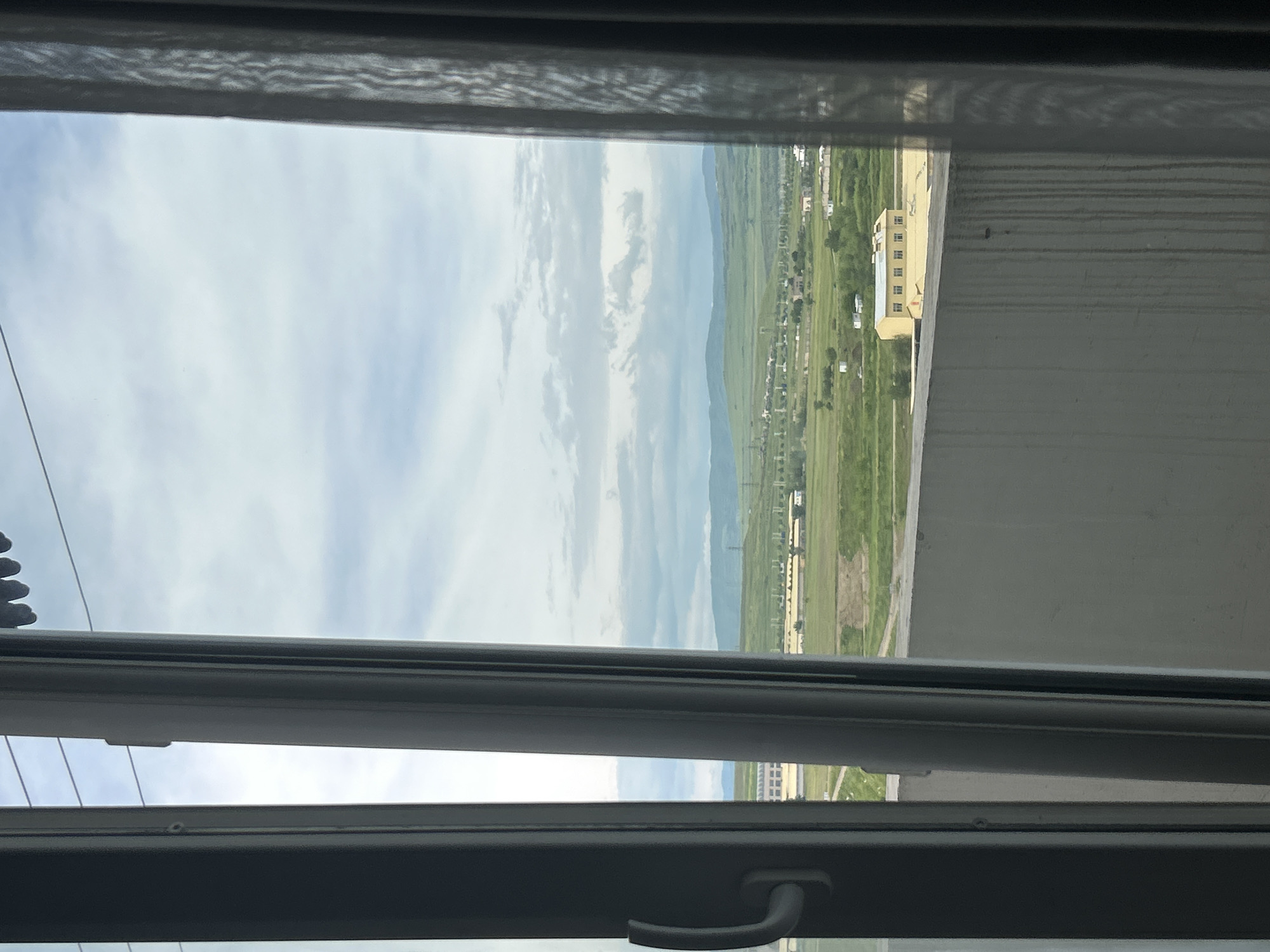 View from a window in Tbilisi, Georgia. The window is partially open, revealing a landscape with a clear sky and distant mountains. A tall building is visible in the mid-ground next to a paved area. There is a town in the background.