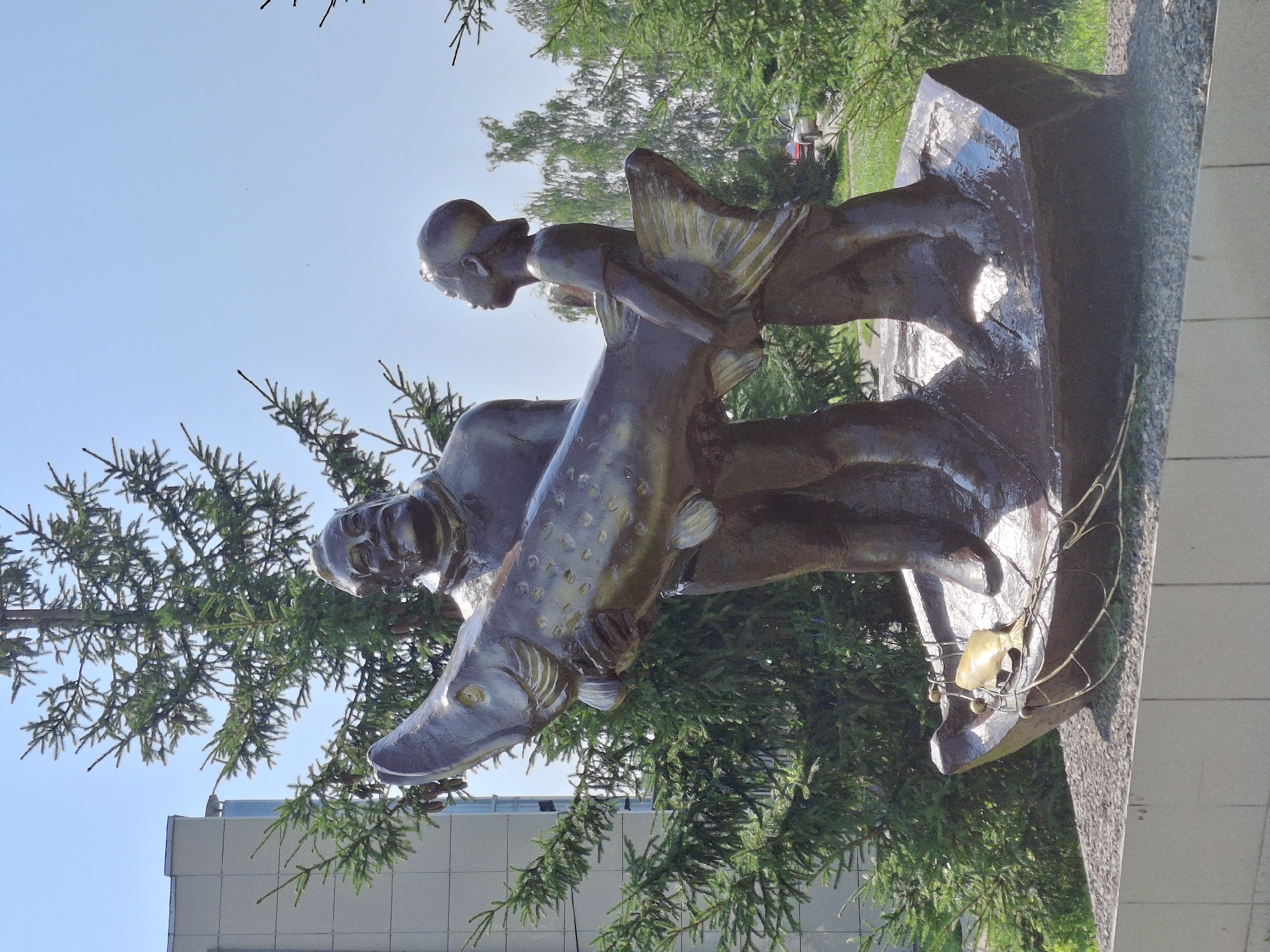 This is a low-angle, full shot of a bronze sculpture depicting a man and a boy in a boat with a large fish. The man, who appears to be older, is on the left, holding the fish's head and chest. The boy, smaller and wearing a baseball cap, is on the right, holding the fish's tail. The fish is depicted as very large, almost larger than the boy. The sculpture is situated on a raised, boat-shaped base that rests on a pebbled surface. The background features evergreen trees, a light-colored building on the left, and a clear blue sky. The sculpture is located in Elovo, Russia, and appears to be a monument celebrating fishing. The sunlight casts reflections on the wet-looking surface of the sculpture.