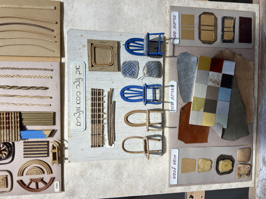 A wall-mounted educational or craft exhibit in Yokneam Ilit, Israel, systematically displays materials, miniature furniture components, and samples illustrating chair and upholstery design, material science, and craftsmanship. The display features three distinct, vertically stacked panels against a light-colored, textured wall.
The **top panel** showcases raw materials and construction techniques. It includes light-toned wooden or reed elements (coiled, curved pieces, segmented slats, a small wooden sleigh model), braided and twisted cords, thin linear strips, and a section of woven material with blue elements. A Hebrew label "לולר" (Lular) is visible in the lower left.
The **middle panel**, set against a beige fabric-like background, focuses on chair construction. A central white plaque bears the Hebrew title "איך רונר כיסא חפצים," translating to "How to make chair objects" or "Chair object manufacturing process." Above the title are samples of horizontal woven matting and bundled reeds. To the upper right, a square frame with an inserted curved rattan piece appears, possibly a jig. Below these are three miniature chairs: two natural wood or rattan frames without seats or backs, and one fully assembled and painted bright blue. Adjacent are two small textile samples (one blue with cross-stitching, one grey/brown knitted with a needle) resembling miniature cushions or backrests. A fourth blue chair has a grey fabric cushion on its seat.
The **bottom panel**, presented on a light brown board, is dedicated to material swatches and padding samples. The left section, labeled in Hebrew "כסף עשוי" (Keseph Asui - contextually padding/cushioning), displays four yellow/beige foam-like samples. The central section, labeled "חומרי מלאכה" (Chomrei Melakha - Craft Materials), presents an array of swatches, including brown/grey leather, a silver metallic sheet, and a grid of colored squares featuring various hues and textures (e.g., beige, yellow, olive green, blue, black, red, white, and a snakeskin pattern). The right section, labeled "כריכת ספסל" (Krichat Sapsal - Bench Upholstery), exhibits four rectangular material samples in shades of light and darker reddish-brown for upholstery.
No individuals are present. The uniform lighting suggests an indoor, well-illuminated setting typical of a gallery or educational institution.
