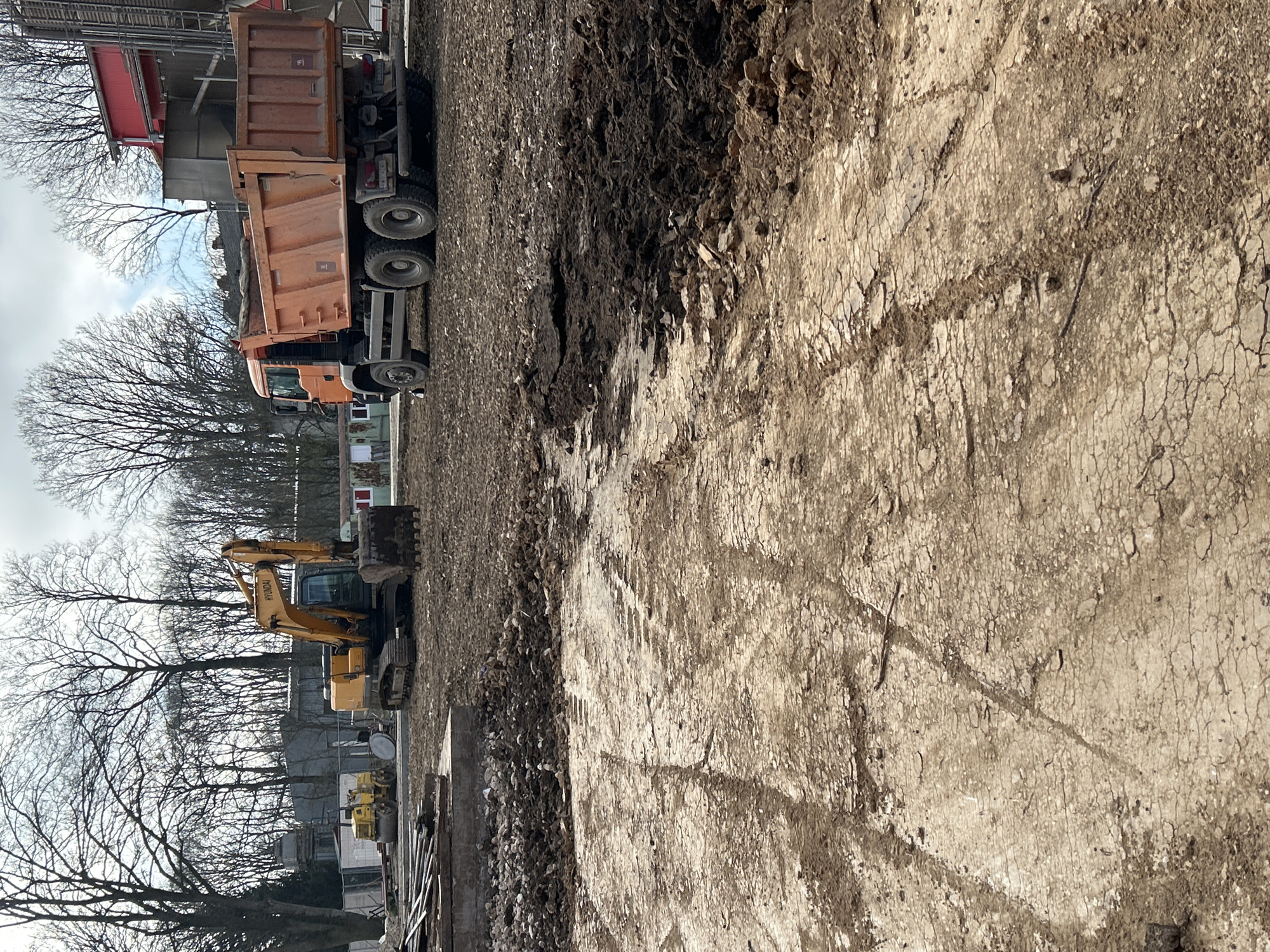 The image depicts a construction site in Donchevo, Bulgaria, under an overcast sky. The foreground consists of heavily disturbed earth, showing a mix of dry, cracked soil and darker, wet mud, with clear tire and track impressions from heavy machinery. In the midground, a yellow Hyundai crawler excavator is parked on the left, its bucket resting on the ground. Further left and behind the excavator, a smaller yellow vibratory roller is also present. To the right, an orange multi-axle dump truck (e.g., MAN TGS) is parked facing away from the viewer, its bed empty. The background features a line of leafless deciduous trees, suggesting late autumn or winter. Several utilitarian structures are visible behind the machinery, including a white construction fence, a small grey building with a red roof, and industrial-like components. No personnel are visible.