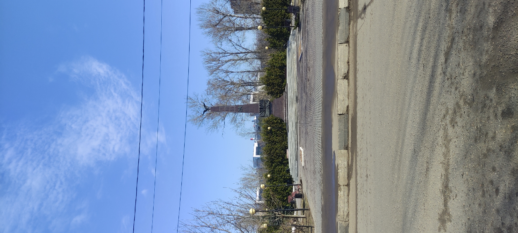 The image captures a street view in Fryazino, Russia, featuring the "Monument to the Fallen Soldiers in the Great Patriotic War 1941-1945". A tall column monument with a bird sculpture atop is centrally positioned. The sky is blue with scattered clouds. Power lines intersect the view from the left side. The right side of the image shows a street or sidewalk and green bushes. Lampposts line the sidewalk. Trees without leaves are also present. A section of a low-rise building is visible in the background.