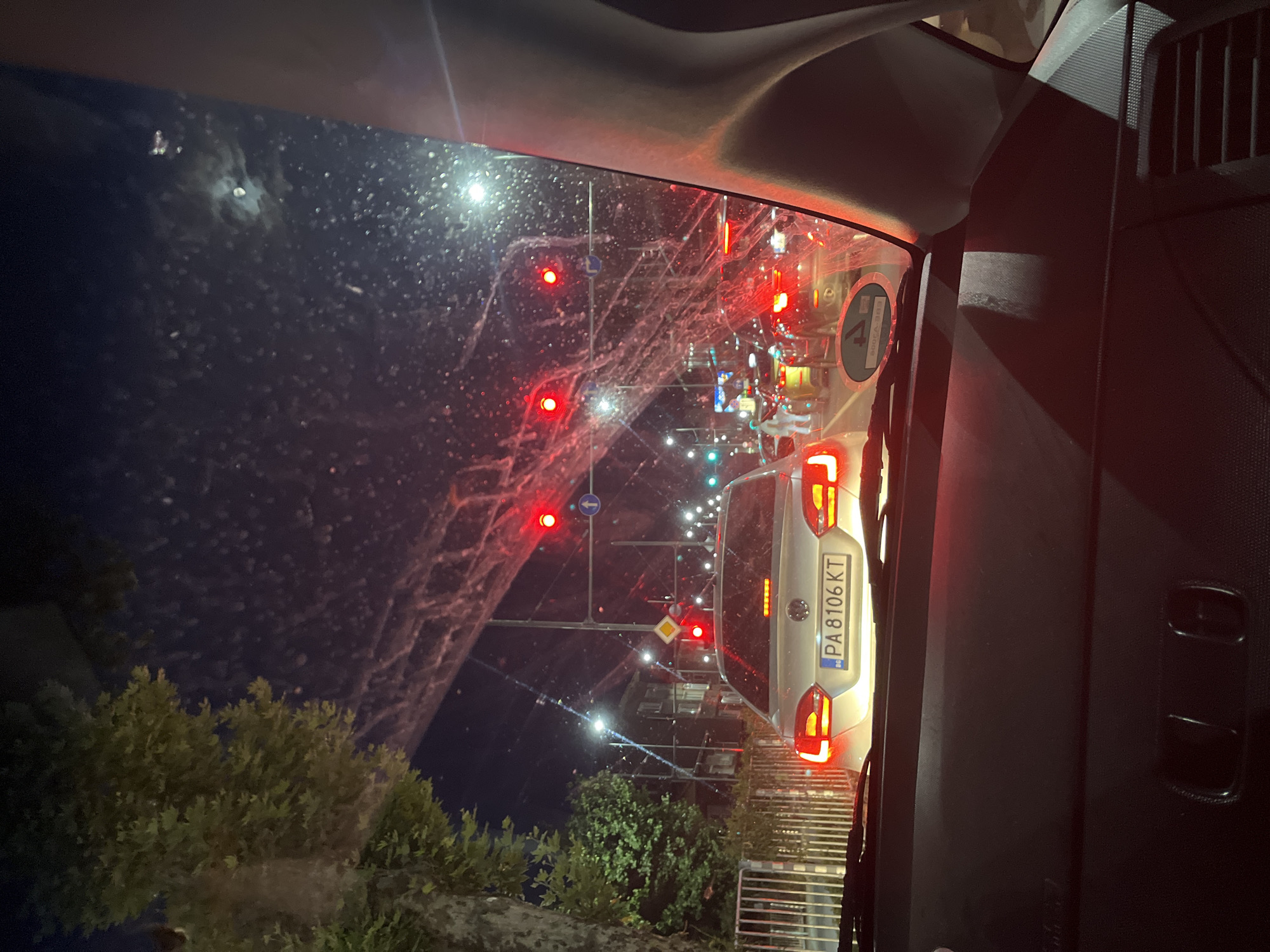 The image depicts a night-time street scene in Plovdiv, Bulgaria, viewed from inside a vehicle. Traffic lights, predominantly red, illuminate the wet windshield, which is streaked with dirt and water droplets. Several cars are stopped in traffic. The license plate of the car directly in front reads "PA 8106 KT". On the left side of the road, a tree with green foliage is visible, along with a metal fence. Buildings and streetlights are present in the background. The moon is faintly visible in the dark sky.