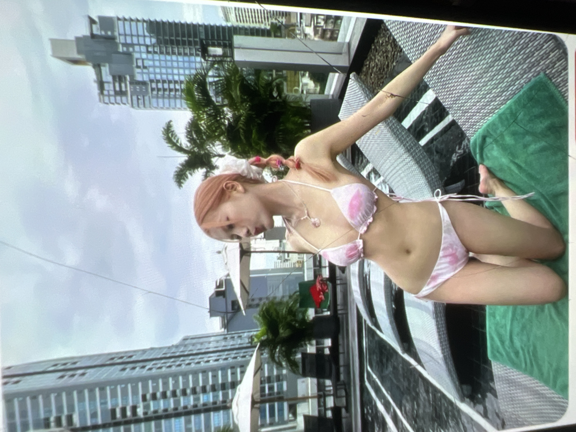 The image shows a young woman in a pink and white patterned bikini posing on a rooftop pool deck. She has blonde hair styled in pigtails and is wearing a necklace. In the background, there are tall buildings, palm trees, and lounge chairs with umbrellas around a pool. The location is identified as Kara-Dıykan, Kyrgyzstan.
