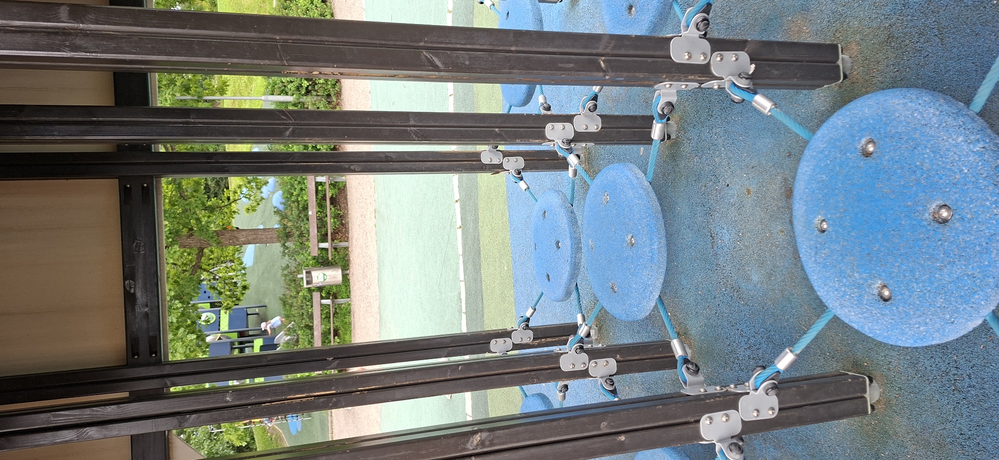 The image shows a close-up view of a blue circular play structure with metal fixtures and blue ropes, set against the backdrop of a playground. In the background, there is a green surface, trees, and two park benches, suggesting an outdoor public space. A child in a light blue outfit and pink hat is visible in the middle ground, near a trash can. The architectural elements of the play structure, such as the dark wooden posts and metal brackets, are prominent in the foreground. The overall scene depicts a children's play area within a park setting. The location is identified as Lahti, Finland.
