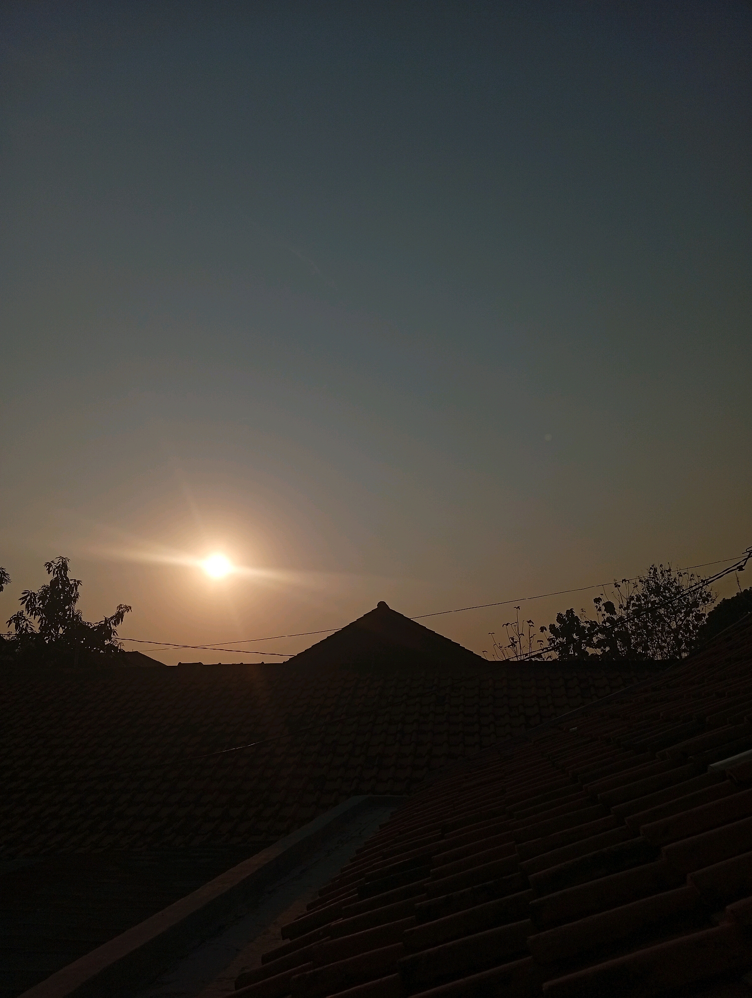 A low-angle shot captures the silhouettes of tiled rooftops against a muted, gradient sky transitioning from a pale blue-grey at the top to a warmer, hazy yellow-orange near the horizon. The sun, a bright orb with visible lens flare, is positioned centrally in the frame, slightly to the left, casting a warm glow and sharp horizontal rays. Bare branches of a tree are silhouetted against the sky to the left of the sun, and another dense cluster of trees with leaves is visible on the right. Thin electrical wires stretch horizontally across the scene, connecting unseen points and further defining the space between the sky and the rooftops. The rooftops themselves are dark and indistinct, their tile patterns visible but without significant detail due to the low light. The overall scene suggests either a sunrise or sunset viewed from a residential area. The image provides no specific clues about the precise location within Kebandingan, Indonesia, beyond it being an area with residential rooftops and vegetation. There are no people or discernable actions visible in the image.
