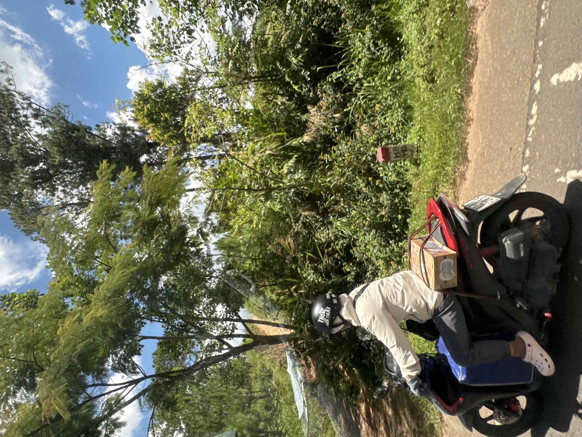 A person rides a scooter on a dirt road lined with lush green foliage and trees under a bright blue sky with scattered clouds. The rider is wearing a light-colored jacket, dark pants, a white helmet with black lettering, and white Crocs. A blue bin is attached to the front of the scooter, and a small cardboard box is secured to the rear rack. The environment suggests a rural or semi-rural setting within Thôn 6, Vietnam, characterized by dense vegetation and a natural landscape. A red and white marker post with the inscription "H8 110" is visible on the right side of the road, indicating a location marker. The road itself appears to be unpaved, with patches of sunlight and shadow.