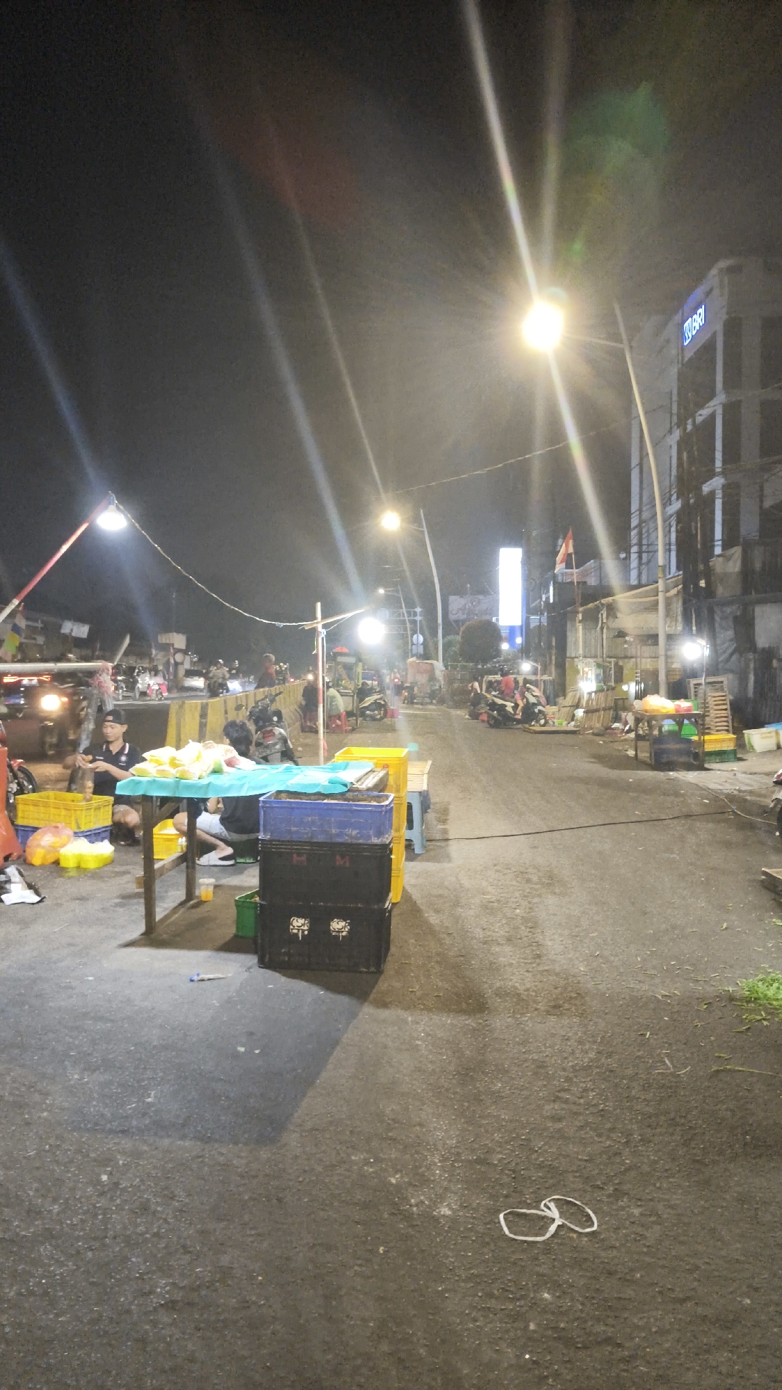 This is a street scene at night in South Jakarta, Indonesia. A group of people are gathered around tables laden with yellow produce, likely fruits or vegetables, under the glare of streetlights. Several motorcycles are parked nearby, and some individuals are interacting with the produce. The street is paved and appears to be an active marketplace. A building with a visible "BRI" logo is visible in the background, indicating a commercial area. The lighting creates strong highlights and shadows, typical of a night scene with artificial illumination. The ground is wet in some areas, suggesting recent rain or ongoing street cleaning.