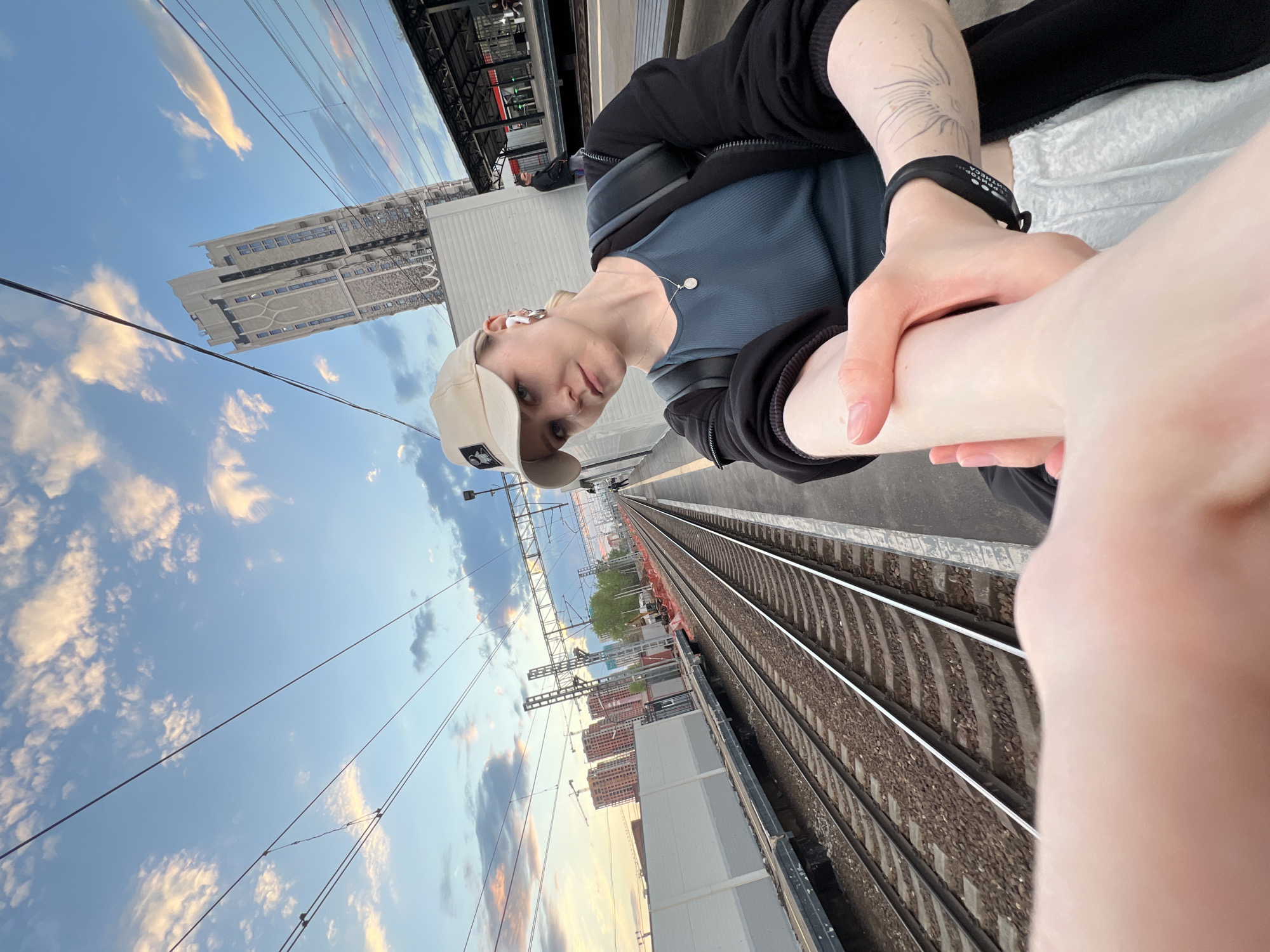 A selfie taken on a train platform in Khimki, Russia, shows a fair-skinned person with short, light hair wearing a light-colored baseball cap, a blue top, and a black jacket. The person is holding a pale forearm. The railway tracks stretch into the distance. A modern building is visible in the background to the left, and other buildings under construction are further down the tracks. The sky is blue with scattered clouds. Overhead power lines are also present.