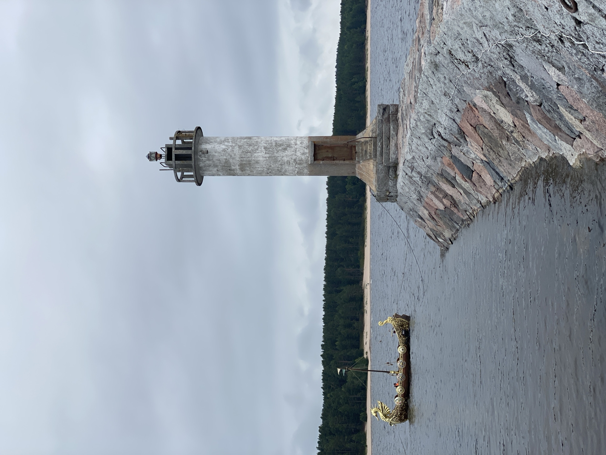 A Viking ship with dragon prows is moored in the water, with a lighthouse and a stone pier extending into the sea. The lighthouse is cylindrical and made of concrete, with a weathered finish and a door at its base. The pier is constructed from large, irregular stones, with a white rope strung along its length. In the background, a dense green forest borders a sandy beach. The sky is overcast and cloudy. The location is the city of Motornoe, Russia.