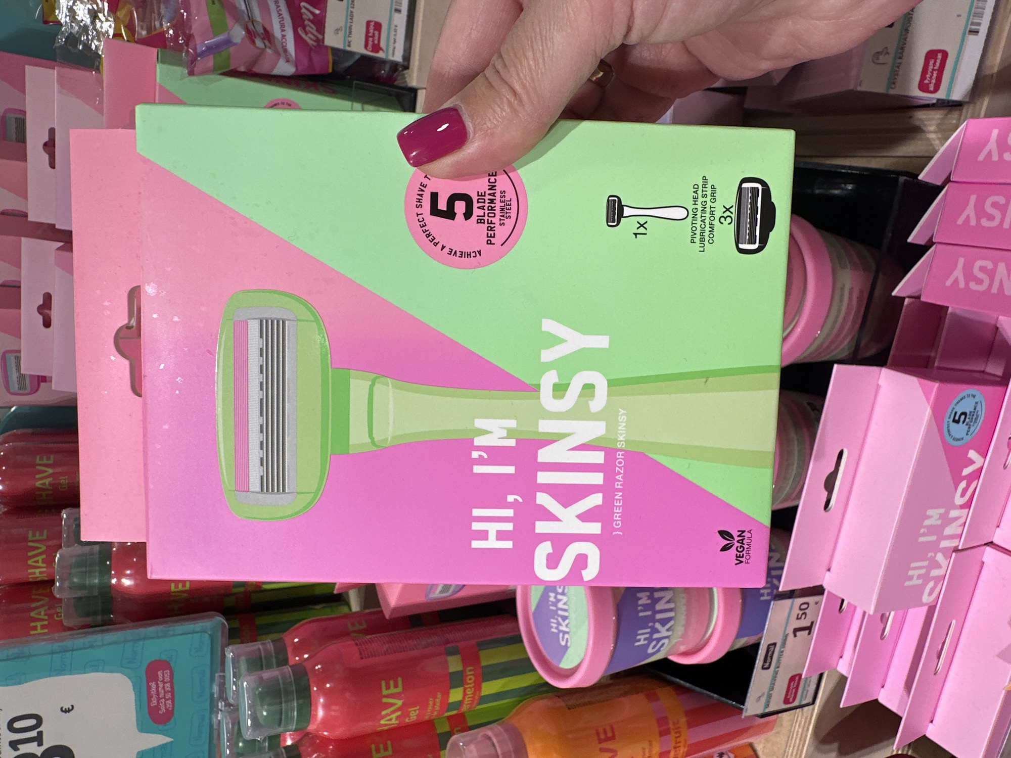 An eye-level, medium shot captures a retail display of "Hi, I'm Skinsy" razors in a store. The primary focus is on a bright pink and green cardboard box showcasing a lime green razor with a pink handle. The box features prominent text reading "HI, I'M SKINSY" in white lettering, along with descriptive details about the product's features, including "5 BLADE PERFORMANCE," "STAINLESS STEEL," and a "VEGAN FORMULA" logo.
To the left of the main box, several pink and red aerosol cans with green caps are visible, some displaying "HAVE" text and what appears to be "WATERMELON" and "FRUIT" labels. A few small pink tubs with lids are stacked below, also showing "HI, I'M SKINS" branding.
Below the main display, rows of smaller pink boxes with the "HI, I'M SKINSY" logo are neatly arranged. A price tag showing "1.50" is partially visible in the lower left.
The background consists of more retail shelving with a variety of personal care products, including more aerosol cans and packaged items. The lighting is bright and even, typical of a store environment. The scene is static, with no discernible movement or interaction depicted. The origin of the image is unknown, as is the specific city and country.