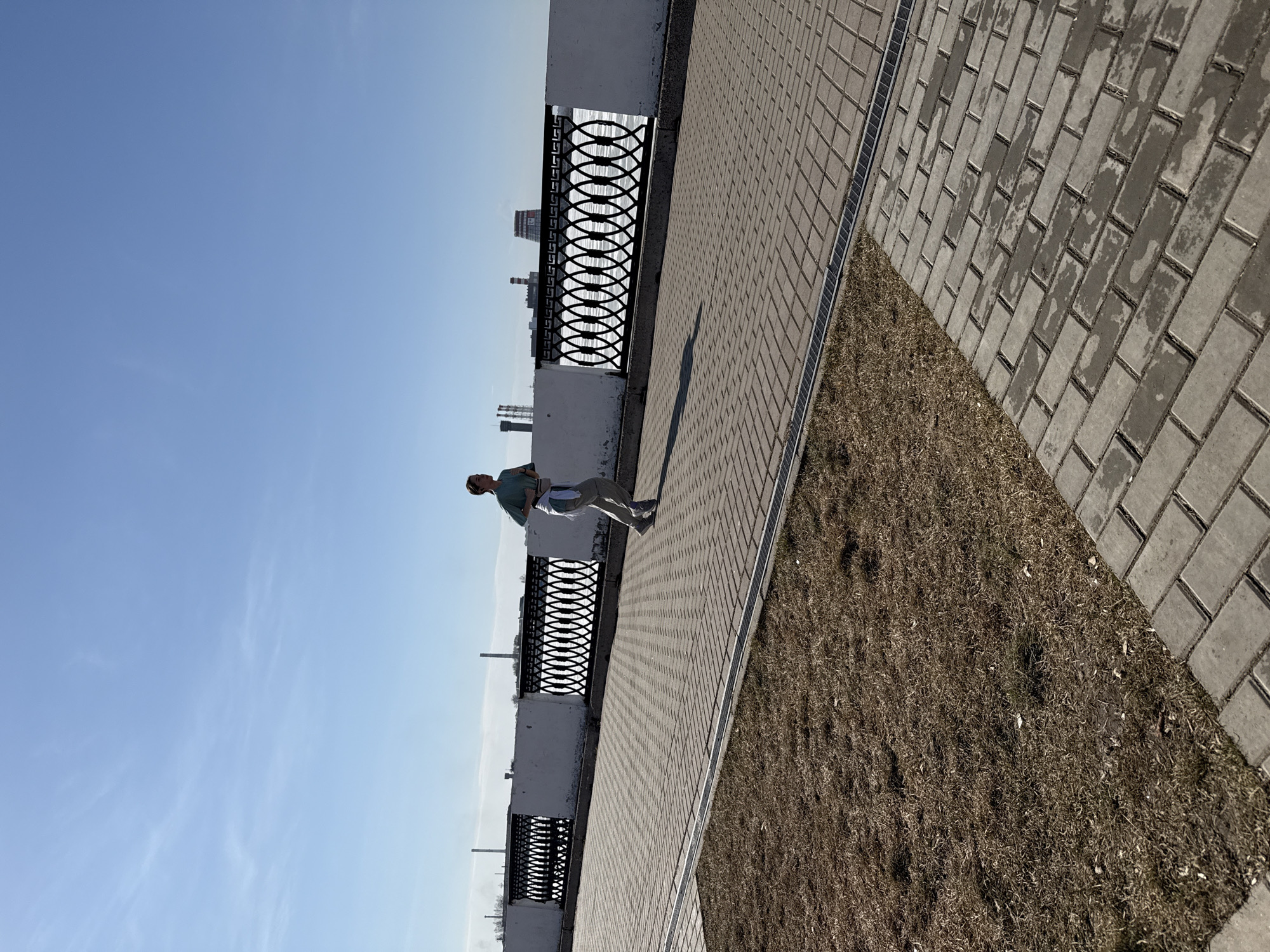 A person is seen on a paved walkway in Izhevsk, Russia, next to a decorative wrought-iron fence and a grassy area. The walkway is made of rectangular paving stones. The person appears to be holding something, perhaps papers or a small bag. In the background, there are structures that suggest a city skyline. The sky is clear and blue.