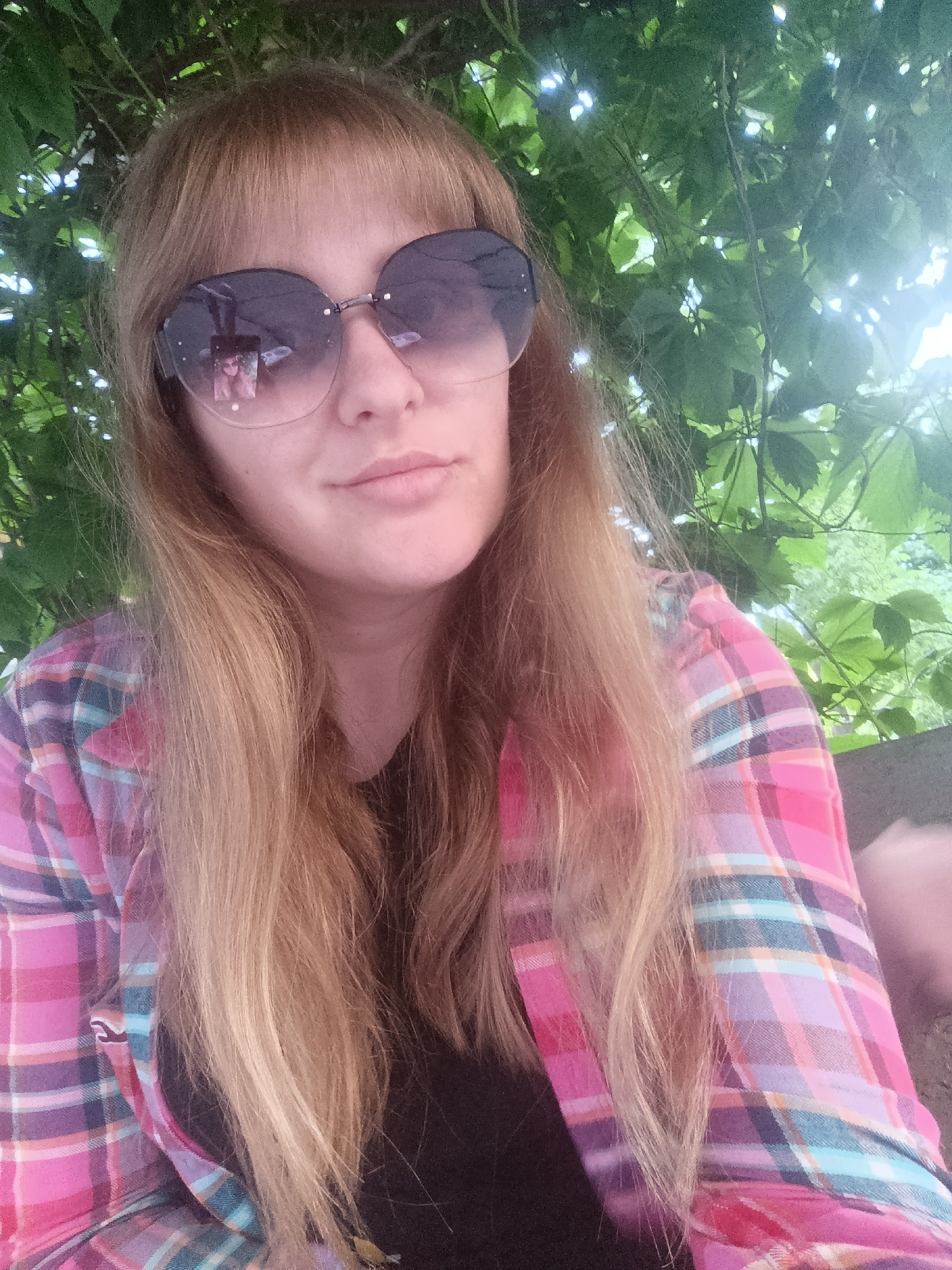The image is a close-up, outdoor selfie of a woman. She has fair skin, bangs, and long, straight, light brown hair. She is wearing large, dark-lensed sunglasses that reflect a small image of her face. Her attire consists of a pink, purple, and teal plaid shirt over a dark t-shirt.
The background is filled with lush green foliage and appears to be dappled with sunlight, suggesting an outdoor setting, possibly a park or garden, during daytime. The light is bright, creating a slight overexposure in some areas. The woman's expression is neutral, with her lips slightly parted. The overall mood is casual and relaxed. There is no visible text in the image.