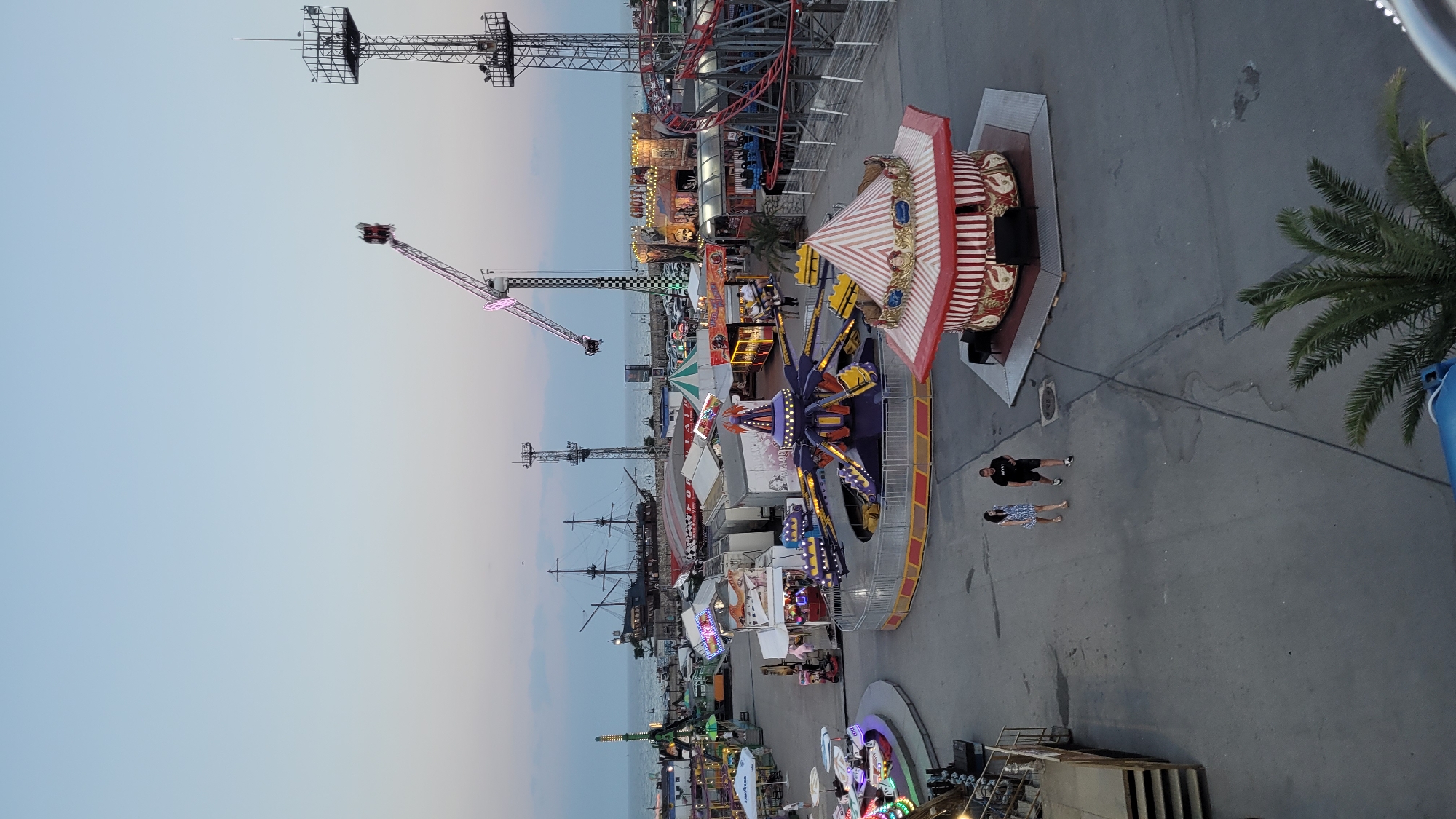The image shows a bustling amusement park scene, likely Varna, Bulgaria, as dusk approaches. The sky is a gradient of soft blues and pinks, indicating either dawn or dusk. Dominating the scene are various amusement park rides, including a towering roller coaster with red tracks, a large swing ride with a crane-like arm, and a circular ride with colorful, illuminated pods. Several smaller rides, characterized by flashing lights and intricate structures, are also visible.
In the foreground, two people, a man in a black t-shirt and shorts, and a woman in a blue and white patterned dress, are walking on the paved ground. Their presence suggests the park is open and active. The park's layout includes walkways, and on the left side of the image, a large pirate ship structure, likely part of a themed attraction, stands prominently.
The atmosphere appears lively, with the colorful lights of the rides illuminating the park. The overall impression is of a vibrant entertainment venue, perhaps nearing the end of a day's operation. There is no discernible text in the image, except for what might be small signs on the rides, which are too blurry to read. The presence of a palm tree on the right adds a touch of coastal ambiance.