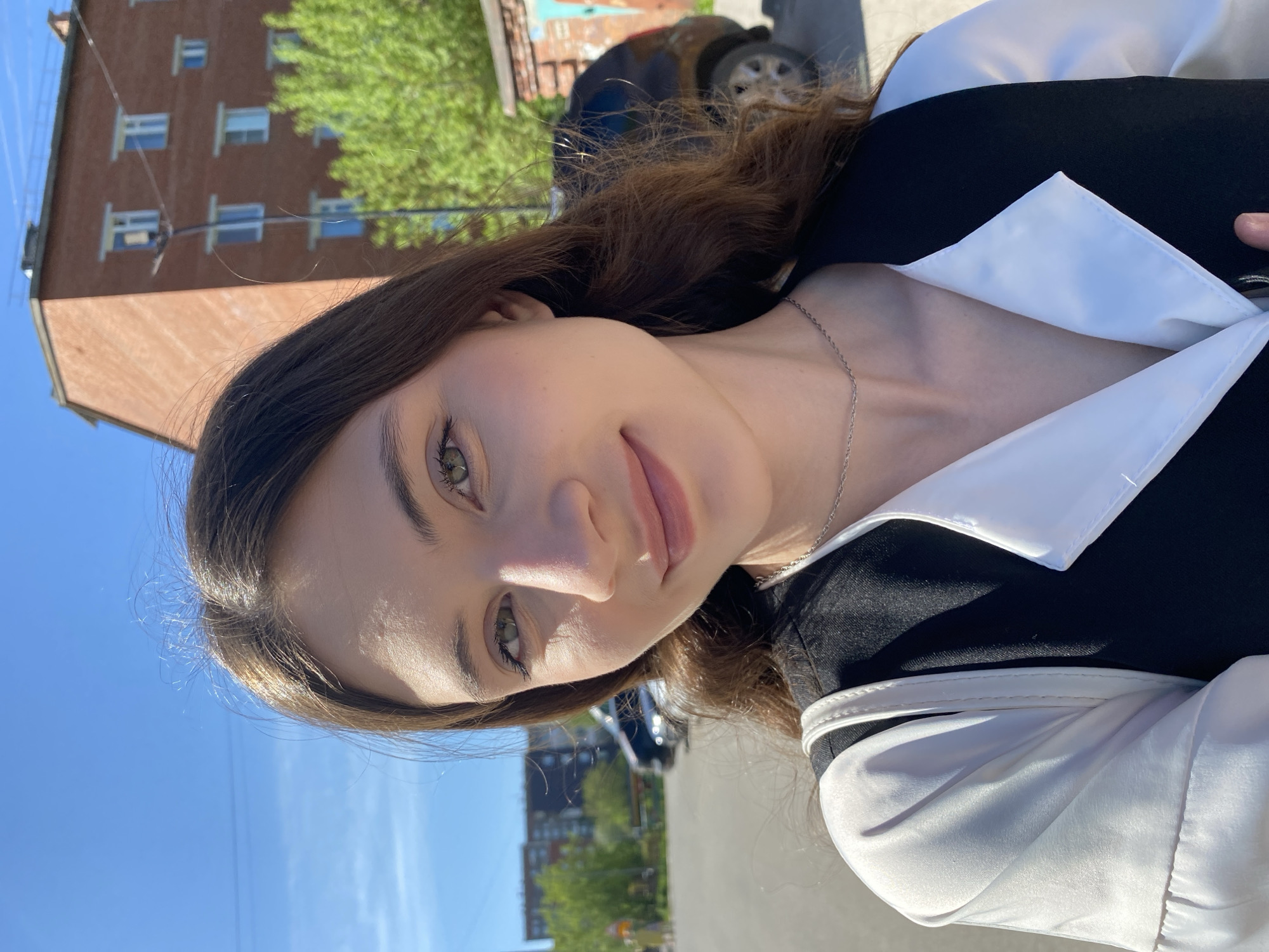 A young woman is posing for a selfie outdoors in Votkinsk, Russia. She has light skin, dark hair, and green eyes, and is wearing a black and white collared shirt. A thin silver necklace is visible around her neck.
In the background, a brick building with multiple windows can be seen on the left. Trees with green leaves are also present, suggesting it is likely spring or summer. A car is parked in the background. The sky is blue and the lighting suggests a sunny day. The photograph is taken from a slightly low angle. The sidewalk is visible.