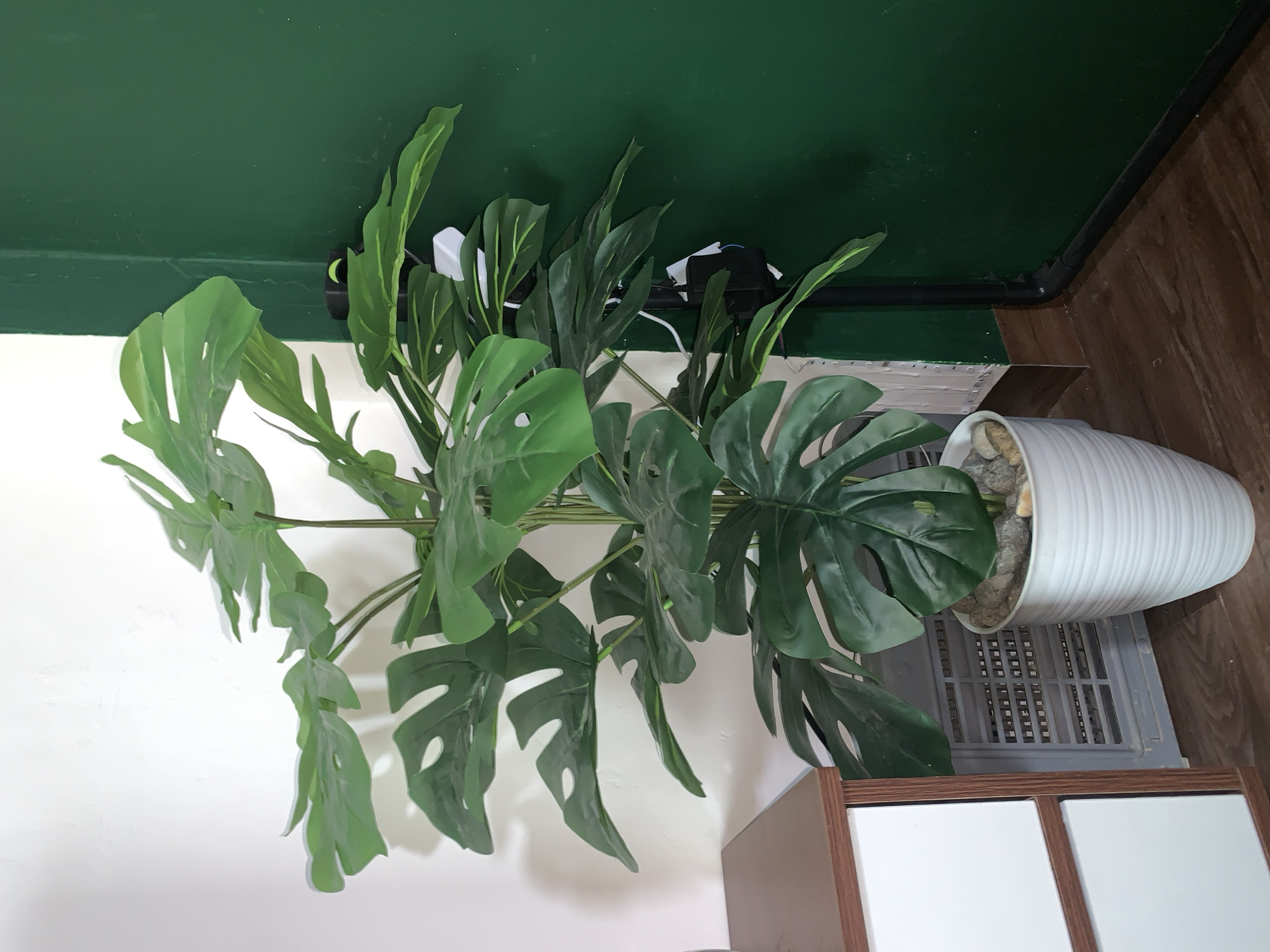 A tall, artificial Monstera plant with large, split green leaves is situated in a white ribbed planter filled with decorative stones. The planter sits on a dark wooden floor. To the left of the plant is a white cabinet with brown trim and two visible drawers. Behind the plant, a grey, grated air vent is visible. To the right of the plant, a section of a wall painted a dark green is present, along with a black electrical conduit and a power strip with multiple plugs. A white wall with some visible texture forms the background on the left side. The scene depicts an indoor setting, likely a room in a residential or commercial space in the city of Kualadua, Indonesia.