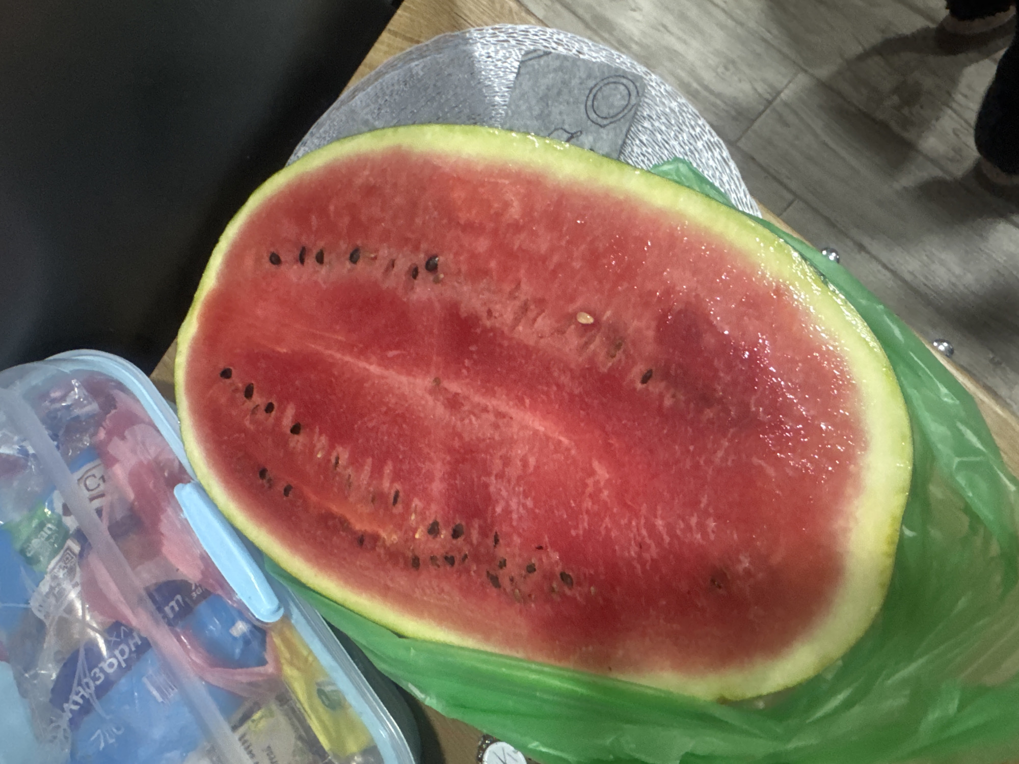 The image depicts a halved watermelon resting on a green plastic bag. The watermelon has a bright red flesh with numerous black seeds visible. The rind is green with a yellowish-white underlayer. Next to the watermelon is a blue plastic container filled with various items. The background includes parts of a grey object and wooden flooring.
I cannot determine any information from this image related to the city of Lehchevo, Bulgaria.