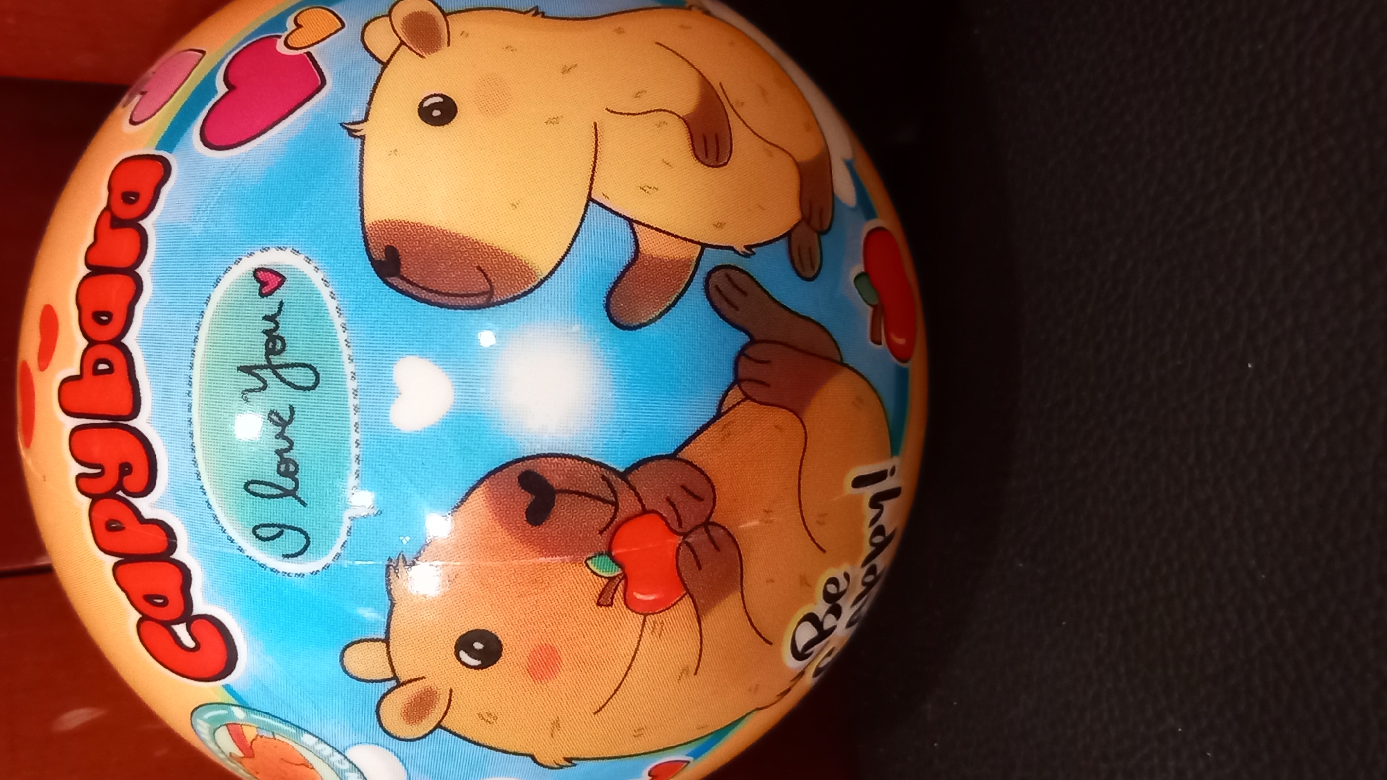 The image is a close-up, slightly angled shot of a spherical object, likely a toy or a decorative ball, with a cartoon design featuring capybaras. The top portion of the ball has the words "Capybara" in large, red, bubbly lettering. Below this, a blue sky with white cartoon clouds serves as the background. Two capybara characters are depicted. The capybara on the left is shown lying down, holding a red apple, and has the phrase "I love You" written in a speech bubble above it, accompanied by a pink heart. Below this capybara, the words "Be happy!" are written. The capybara on the right is sitting and appears to be interacting with the other. Several pink and orange hearts are scattered around the design. The overall color palette is bright and cheerful. The object is placed on a dark, textured surface, possibly a table or fabric. The image does not contain any information or visual cues that would suggest a location in the city of Kos, Greece.
