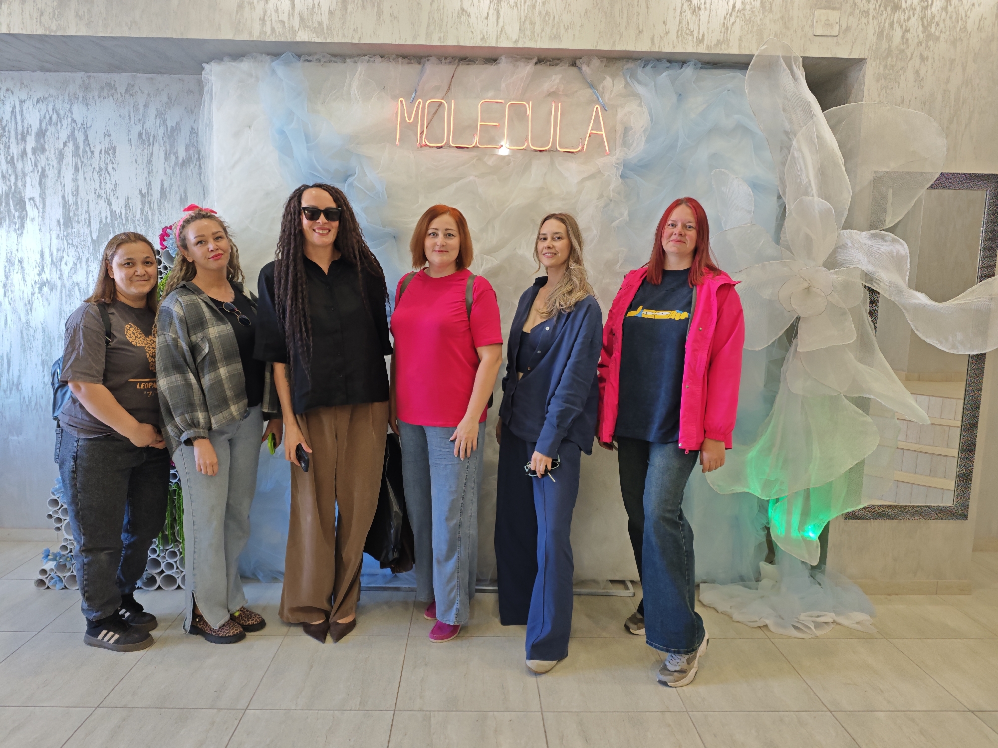 A group of six women are posing for a picture in front of a decorative wall. A neon sign spelling out "MOLECULA" is visible above a background of blue and white tulle-like material. To the right, there is a large, ornate flower-like sculpture crafted from a similar material, partially illuminated with green light, and a mirror. The setting seems to be a shop or event space. The women are dressed in casual attire. From left to right: the first woman wears a grey T-shirt and jeans, with black Adidas sneakers; the second woman wears a flannel shirt and jeans, with animal print shoes and a pink flower hair accessory; the third woman wears a black button up shirt and brown pants; the fourth woman wears a pink T-shirt and jeans; the fifth woman wears a navy blue jacket and pants; and the last woman wears a blue T-shirt and jeans with a pink jacket. Based on the "MOLECULA" sign, which is a known brand in Russia, and publicly available information about the brand, the location is most likely in the city of Chaikovsky, Russia.