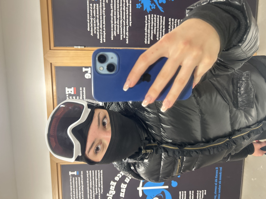 An individual is depicted taking a mirror selfie indoors in Belmont, France. The person wears a black balaclava, white-framed ski goggles with dark lenses pushed onto the forehead, and a shiny black puffer jacket. The jacket features a gold-colored zipper with a black ruffled fabric trim along its length, a zippered pocket on the left chest, and a buttoned flap pocket on the lower right side. The right hand holds a blue smartphone, identifiable by its triple-lens camera module, obscuring part of the individual's face and upper torso. The nails on the visible fingers are manicured with white tips. The background consists of large, dark-colored panels or posters with white, reversed French text and light blue graphic elements, separated by vertical wooden frames. The environment suggests a public indoor space, possibly a visitor center or lodge.