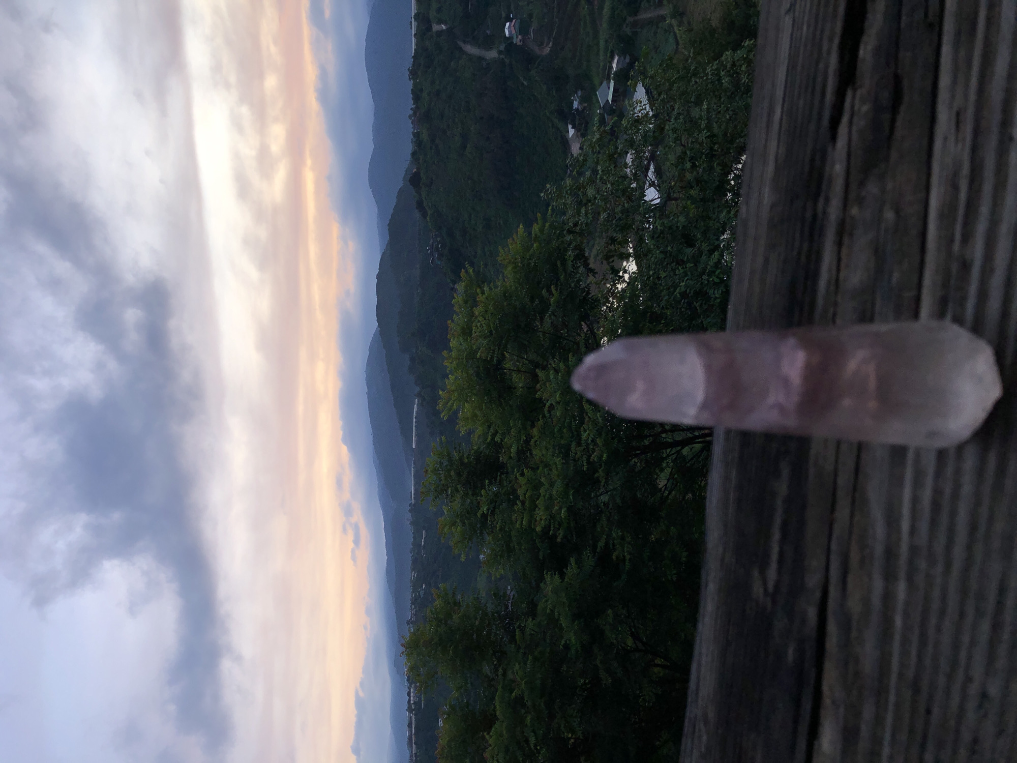 An upright, translucent pink crystal is in the foreground, positioned in the center of the frame. It rests on a dark, weathered wooden surface, likely a railing or table. Behind the crystal, a verdant landscape unfolds. Lush green trees fill the middle ground, interspersed with hints of buildings and roads winding through the hills. In the distance, layers of mist-shrouded mountains fade into the horizon, beneath a soft, twilight sky painted with hues of pink, orange, and pale blue, characteristic of a sunset or sunrise. The image evokes a sense of peaceful observation of nature. The precise location within Thôn 3, Vietnam, is not discernible from the visual cues alone.