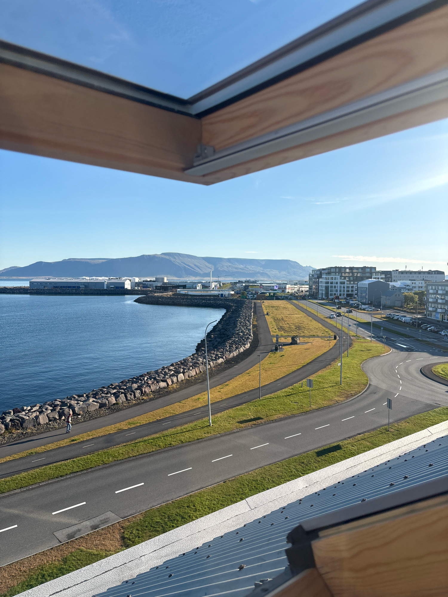 The image captures a panoramic view of Reykjavik, Iceland, from an elevated perspective, likely a building or a skylight. The foreground features the edge of a wooden-framed skylight, with the blue sky visible through it. Below, a corrugated metal roof extends into the frame.
The middle ground reveals a coastal scene with a dark, rocky embankment bordering a calm, blue body of water. A paved path runs along the embankment. Further inland, a road curves through a grassy area, with streetlights and street signs visible. Several cars are parked along the roadside and driving on the road. In the distance, a row of modern buildings lines the coast, interspersed with industrial structures. A lone figure, appearing to be a pedestrian, walks along the path by the water.
The background is dominated by a hazy, mountainous landscape under a clear, bright blue sky with the sun faintly visible. The overall impression is one of a bright, sunny day in a coastal city.
