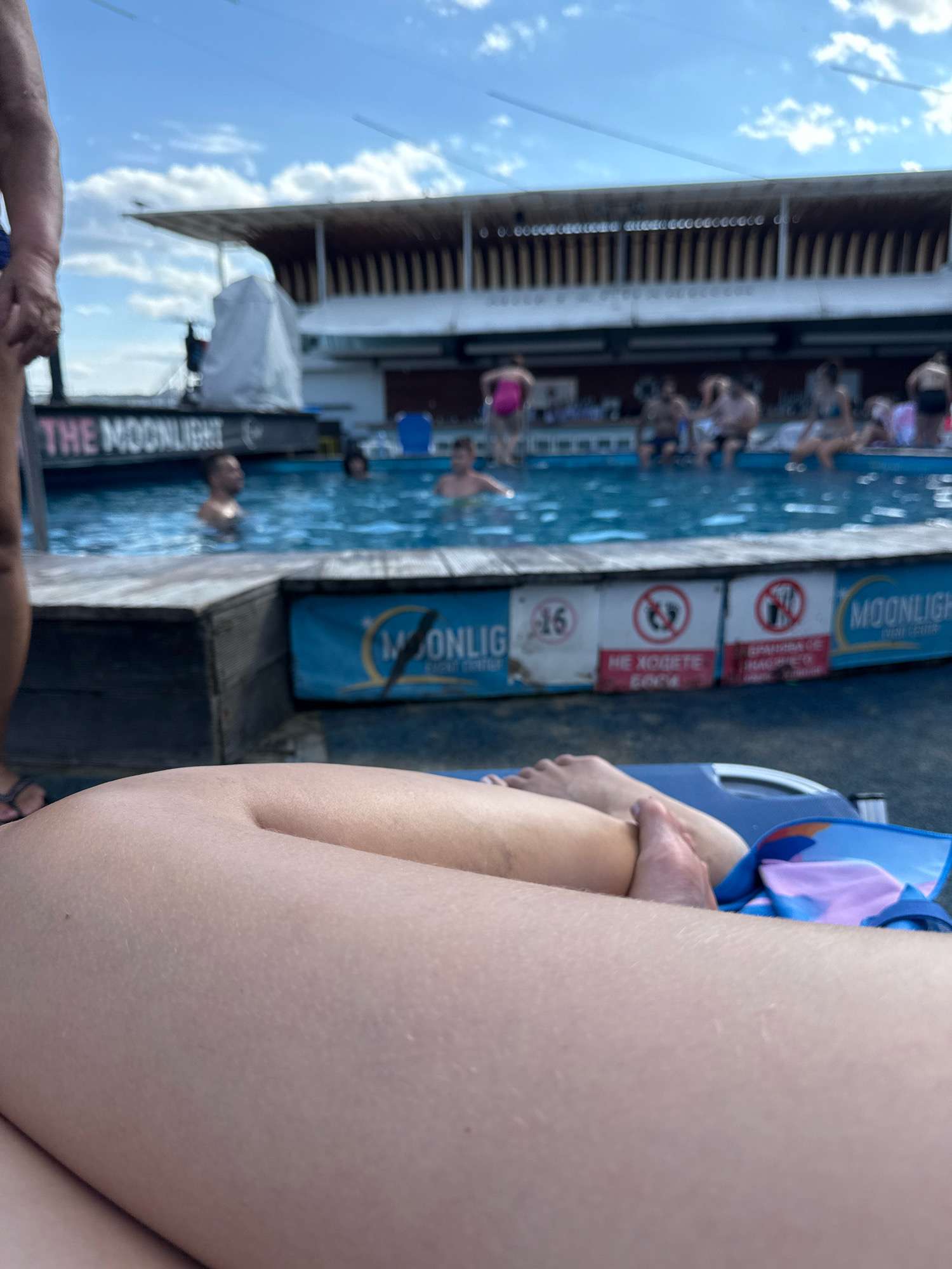 A first-person perspective shot shows a person lying down on a blue lounge chair with a towel draped over it. The person's legs are in the foreground, out of focus. In the background, a swimming pool is visible, with several people swimming or sitting on the edge. Behind the pool, a building with a large awning and wooden accents stands under a bright blue sky with scattered clouds. Signs are attached to the edge of the pool, including one that says "THE MOONLIGHT".