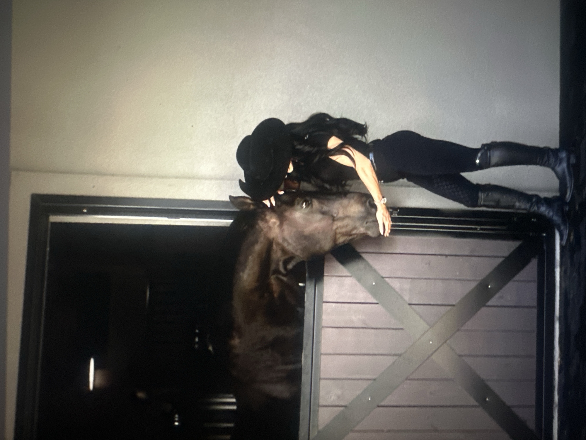 A woman in a black cowboy hat, black top, black patterned leggings, and black boots stands next to a horse in a stable. The horse is dark brown and its head is extended out of the stable door. The woman is reaching out to touch the horse's face. The stable door is made of dark wood with crisscrossing beams. The background is a light gray wall. The scene takes place in a stable, possibly in a rural or suburban setting, not in the city of Unknown, Unknown.