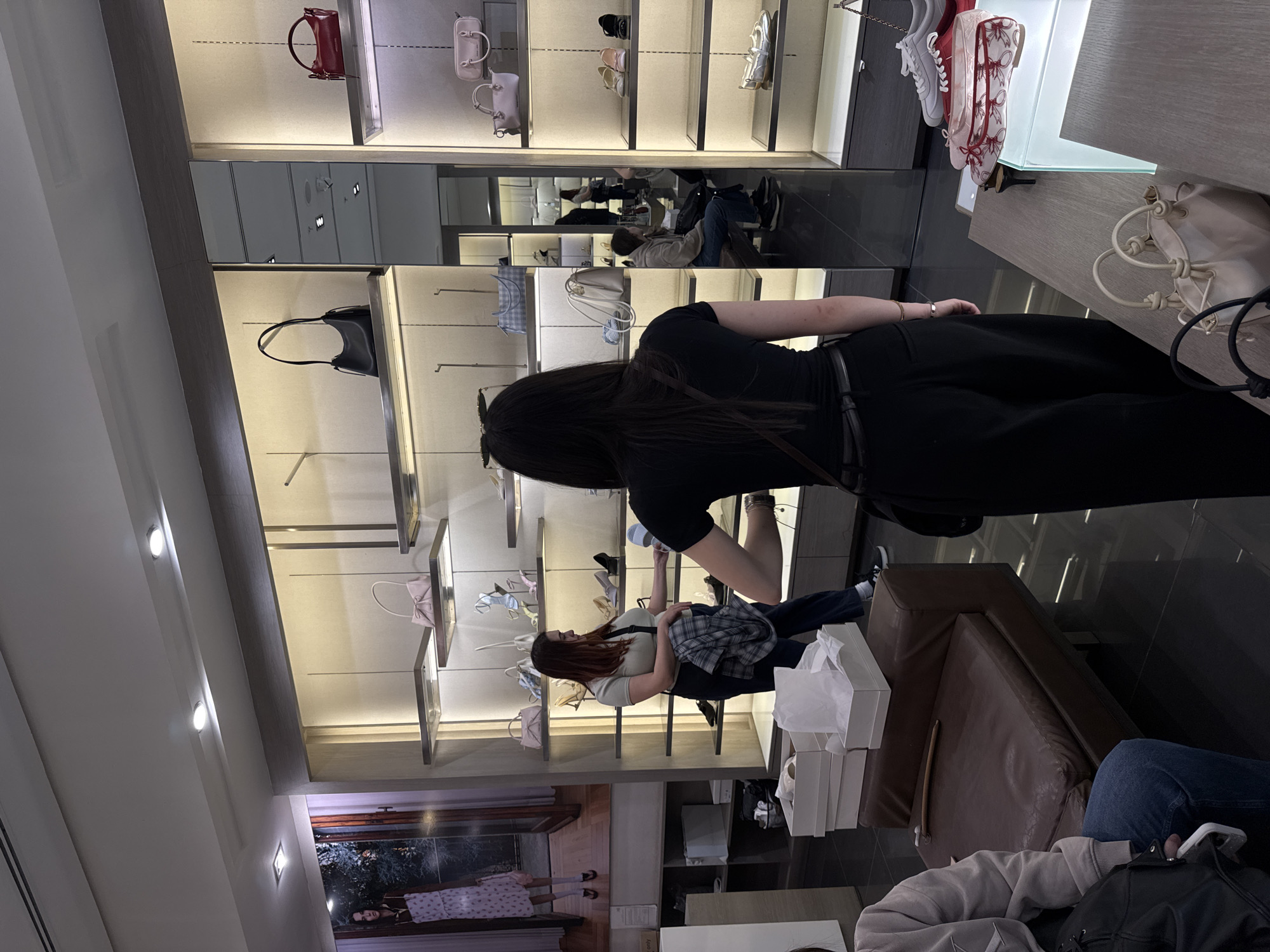 The image shows the interior of a store, possibly a high-end retail outlet, with shelves displaying handbags and shoes. Several people are present: one woman in a black dress is standing and partially obscuring the view, while another woman in a light-colored top and plaid shirt is sitting and appears to be examining items on the shelves. Shopping bags are stacked on a brown sofa near the seating area. In the background, there are more shoppers and a reflection suggesting a mirrored wall. A storefront picture of a model is on the lower left. There is no verifiable evidence to determine that this image was taken in Yerevan, Armenia. Therefore, the city is unknown.