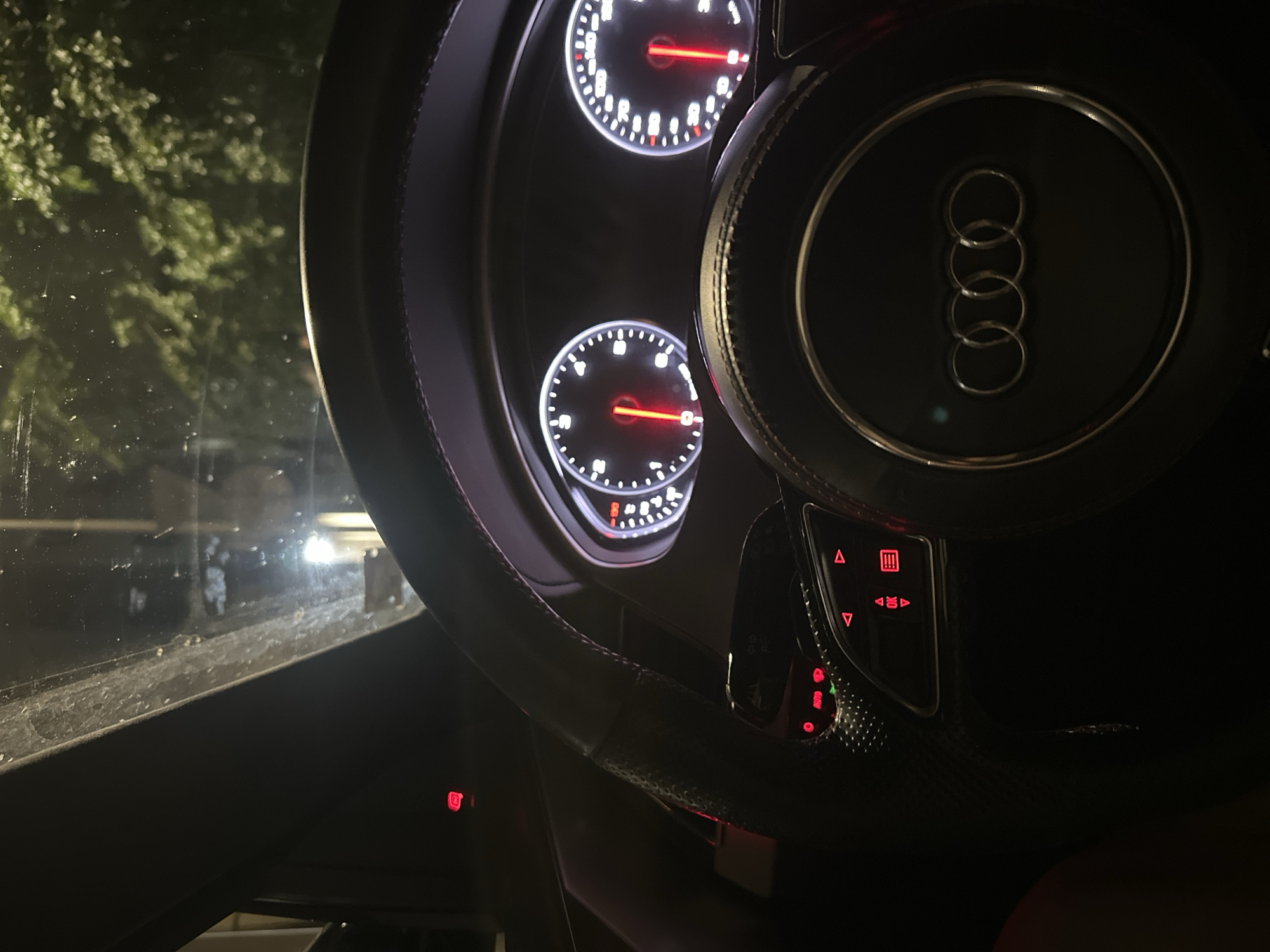 The image shows the interior of a car, specifically the dashboard and steering wheel. The steering wheel has the Audi logo in the center. The dashboard features two circular gauges, likely the speedometer and tachometer, illuminated with red needles. There are also various buttons and controls on the steering wheel. Red indicator lights are visible on the dashboard and steering column. The windshield is dirty and reflects some light, showing glimpses of trees and an outdoor environment at night. It is not possible to determine the specific location in the city of Ruse, Bulgaria from this image alone.