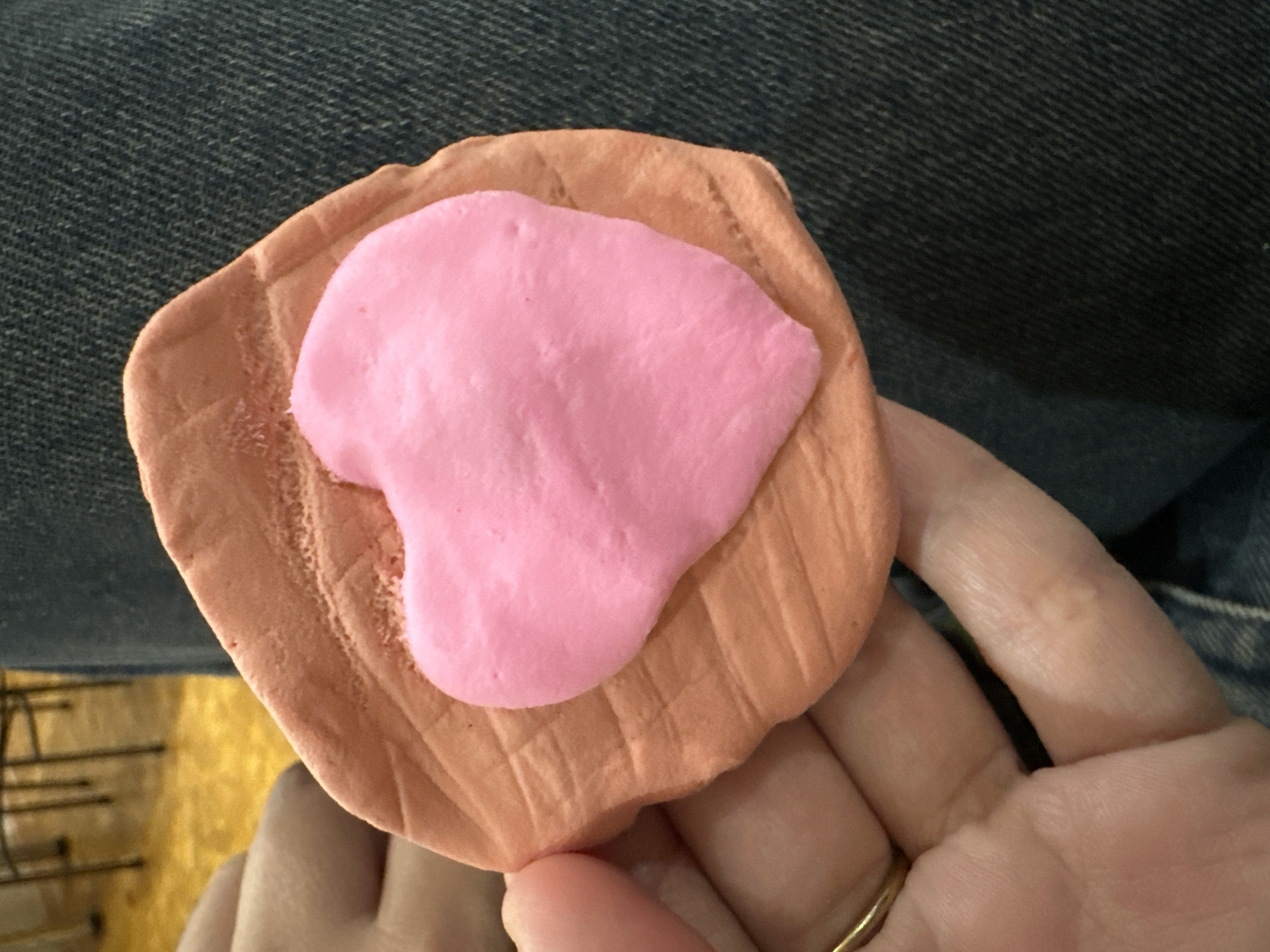 A close-up shot shows a person's hands holding a handmade object, likely crafted from modeling clay or playdough. The primary object consists of a light brown, roughly rectangular or oval base with a distinct textured, ridged surface. Layered on top of this base is a smaller, irregularly shaped blob of bright pink material, resembling an abstract form or a stylized heart.
The person's left hand, with a gold-colored ring on the ring finger, supports the object. The background primarily features the person's lap, covered in blue denim fabric, indicating a casual setting. In the lower left portion of the image, a blurred yellowish area with dark lines suggests an indoor environment, possibly a floor or furniture. The activity depicted is the holding or presentation of this craft item. No specific time of day, weather, or discernible emotions are evident from the image. There is no visible text. The scene is situated in Uva, Russia.