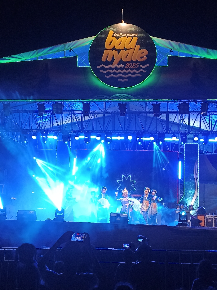 The image depicts a vibrant night scene at an outdoor stage in Kecamatan Pujut, Indonesia, during what appears to be the "Festival Pesona Bau Nyale 2025." A large, circular sign is prominently displayed above the stage, featuring the text "festival pesona" at the top, "bau nyale" in large yellow/orange letters in the center, and "2025" below it, along with wavy lines suggesting water. This sign is encircled by a bright green neon ring, matching other green accent lights on the stage structure.
The stage is brightly illuminated by numerous blue stage lights, casting strong beams through visible haze or smoke, creating a dramatic atmosphere. Overhead, metal trussing supports various lighting fixtures. On stage, four performers are visible, dressed in elaborate, possibly traditional, costumes. One central figure wears a distinctive, ornate golden headpiece. They appear to be engaged in a synchronized performance, likely a cultural or theatrical act.
In the foreground, the silhouettes of an audience can be seen, with several individuals holding up smartphones to record or photograph the event. The time of day is clearly night, characterized by the dark sky and extensive artificial lighting. The scene conveys an energetic and engaging event.