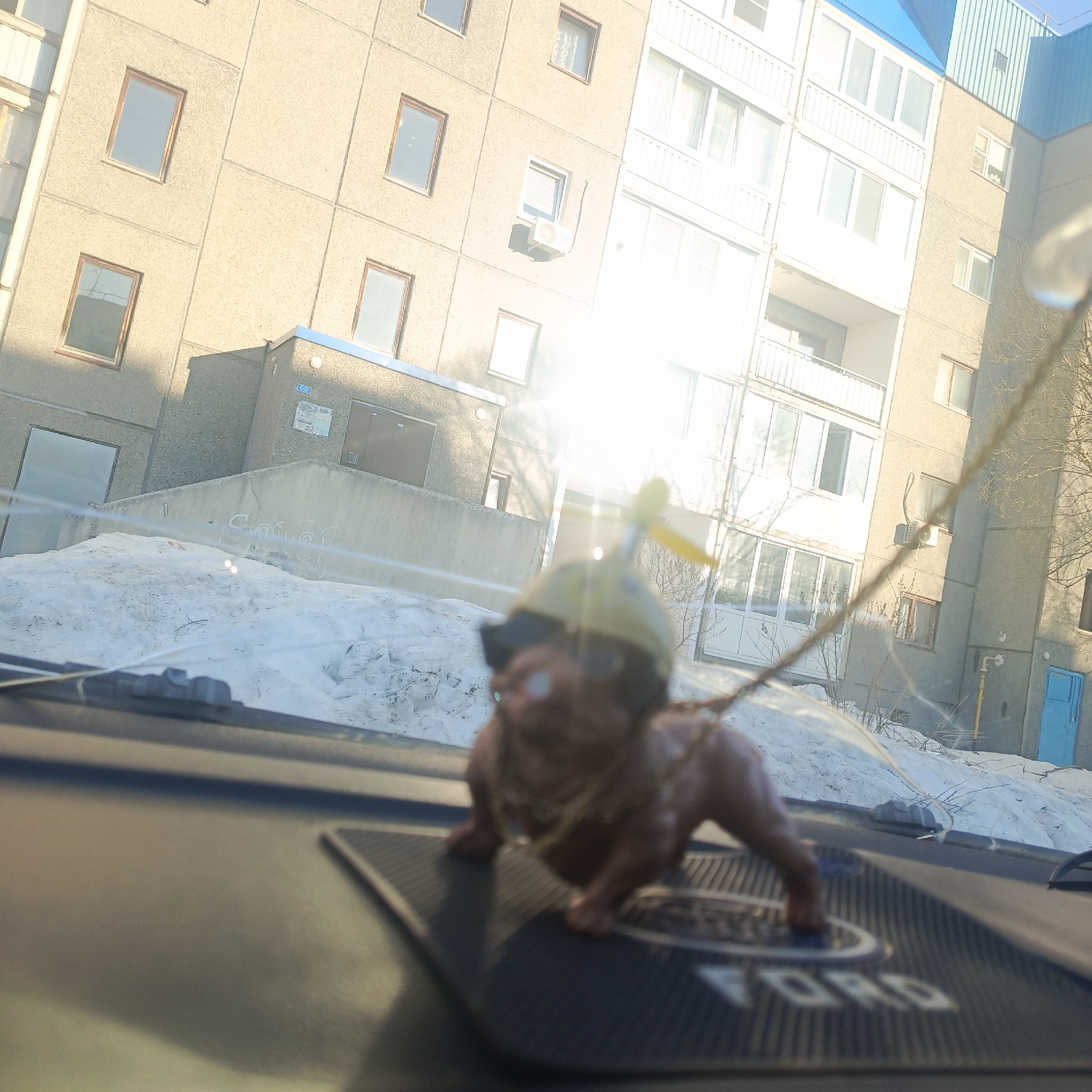 The image, taken from inside a car, shows a doll with a propeller hat and sunglasses on the dashboard. The doll is positioned on a Ford-branded pad. Outside the car, there is snow on the ground, and several concrete apartment buildings are visible. The buildings are typical of urban Soviet-era architecture. One building has a blue roof and blue door, and another features an air conditioning unit attached to the exterior wall. The location is Марковский, Russia.