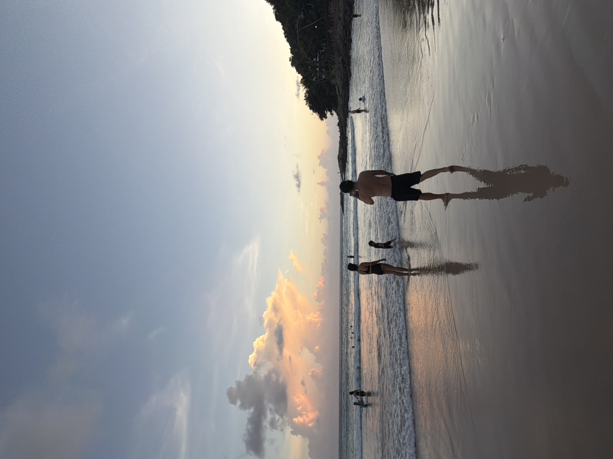 An eye-level, wide shot captures a serene sunset scene at Rawai Beach, Thailand. The sky is a gradient of soft blues and yellows, transitioning into the warm hues of the setting sun. Dramatic, pink-tinged clouds dominate the left side of the frame, contrasting with the clear sky on the right. The ocean, with gentle waves rolling onto the shore, reflects the sky's colors. A sandy beach, wet from the receding tide, stretches across the foreground, mirroring the figures present.
In the foreground, a shirtless man wearing black shorts stands facing away from the camera, holding a phone as if to take a picture or video. His shadow is cast long on the wet sand. To his left, a woman in a black bikini and a child in dark shorts are wading in the shallow water, with another child in dark shorts nearby. Further out in the water, other individuals can be seen enjoying the sea. The right side of the image features a rocky shoreline covered in lush green foliage, characteristic of a tropical landscape. The overall atmosphere is peaceful and idyllic, typical of a beach sunset in Rawai.