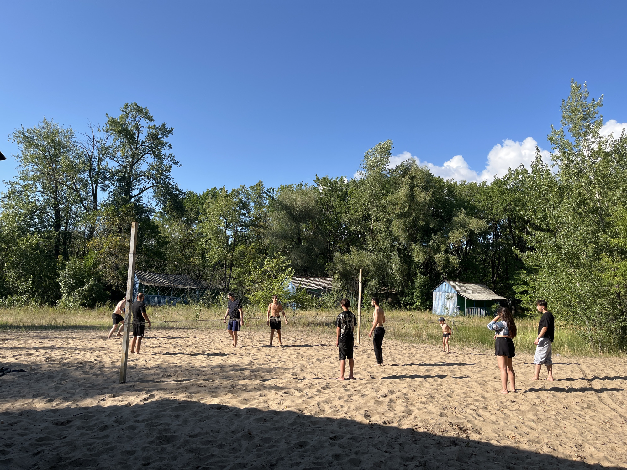 The image depicts a group of people playing volleyball on a sandy court surrounded by trees and vegetation. The setting appears to be a rural or semi-rural area, possibly near a body of water, given the sandy beach-like environment. There are several individuals scattered across the court, some actively engaged in the game, while others appear to be observing or waiting for their turn. A simple volleyball net and posts are visible, defining the playing area. In the background, there are some small structures, possibly sheds or cabins, nestled amongst the trees. The sky is clear and blue, suggesting a sunny day. The overall atmosphere suggests a recreational activity taking place in a natural, outdoor setting. Without further information or context, it is impossible to definitively place this scene within the city of Saratov, Russia, or any specific urban location.