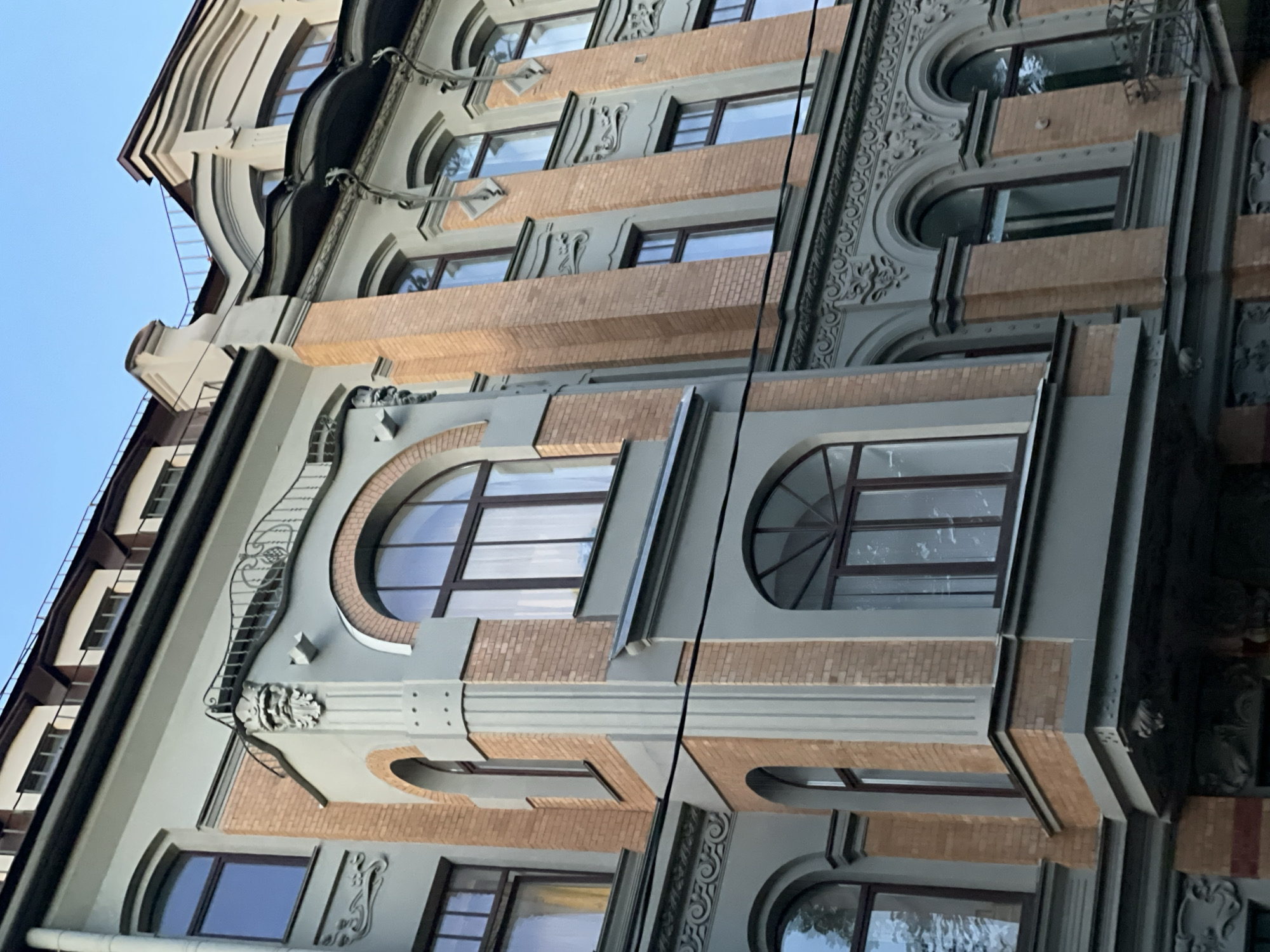 A low-angle medium shot captures the facade of an old, multi-story building in Saint Petersburg, Russia, under a clear blue sky. The building features a decorative architectural style, characterized by contrasting brickwork and painted stucco.
The facade is divided into vertical sections. The lower sections are clad in light brown brick, with some darker red brick accents. Above the brickwork, grey painted stucco is applied, featuring ornate decorative elements, including intricate molding and sculpted faces. Several windows are visible, many with arched tops, framed by darker brown wood or metal. Some windows are clear, reflecting the sky and surrounding buildings, while others are partially obscured by light-colored curtains.
On the left side of the image, a corner section of the building juts out, also combining brick and stucco. This section has a small balcony with a decorative wrought-iron railing, above which is a sculpted face with a crown and beard.
The upper part of the building shows a mansard roof with dormer windows and a dark brown roofline. The overall impression is one of historical grandeur and detailed craftsmanship. The lighting suggests it is daytime, possibly late afternoon given the angle of the light. There are no people or discernible actions taking place in the image.