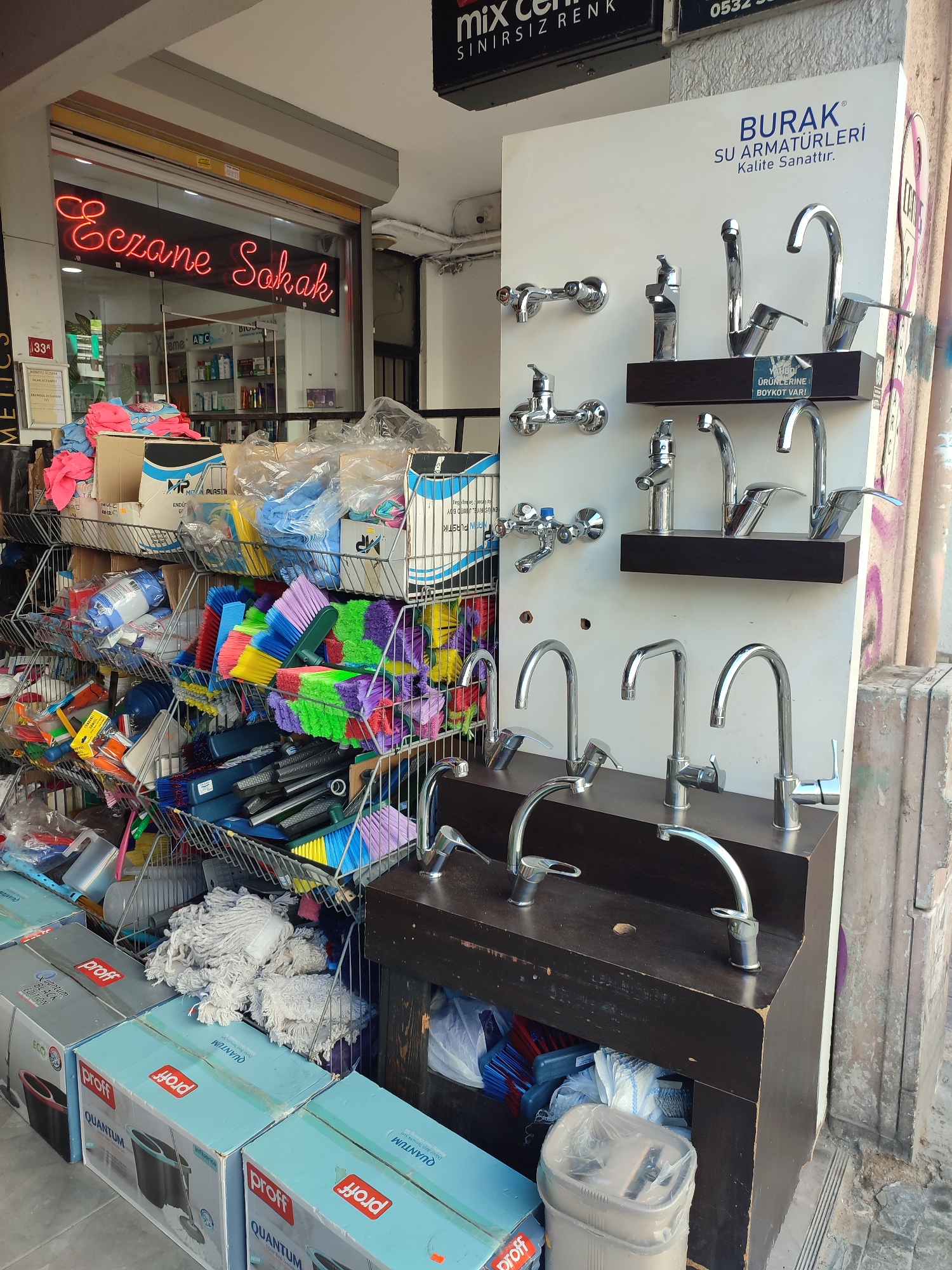 The image shows an outdoor display of plumbing fixtures and cleaning supplies in front of a shop. A neon sign above the entrance reads "Eczane Sakak" in red lettering, which translates to "Sakak Pharmacy." The shop appears to be a retail establishment in Kadıköy, Turkey, given the Turkish signage.
In the foreground, several boxes of cleaning products, specifically "proff QUANTUM" mop sets, are stacked on the ground. Behind these boxes, wire racks are filled with an assortment of colorful cleaning brushes and mops. Further back, more cleaning supplies are visible, including bottles and bags of various products.
To the right of the cleaning supplies is a display of chrome faucets. These faucets are mounted on a white board, with the brand name "BURAK SU ARMATÜRLERI" (Burak Water Fixtures) prominently displayed. The faucets range in style and design, with some featuring high, curved spouts and others with more angular shapes. A dark wood cabinet supports some of these faucets, and below it, more cleaning supplies are visible. The overall scene suggests a busy street-level retail environment with goods displayed for sale to passersby.