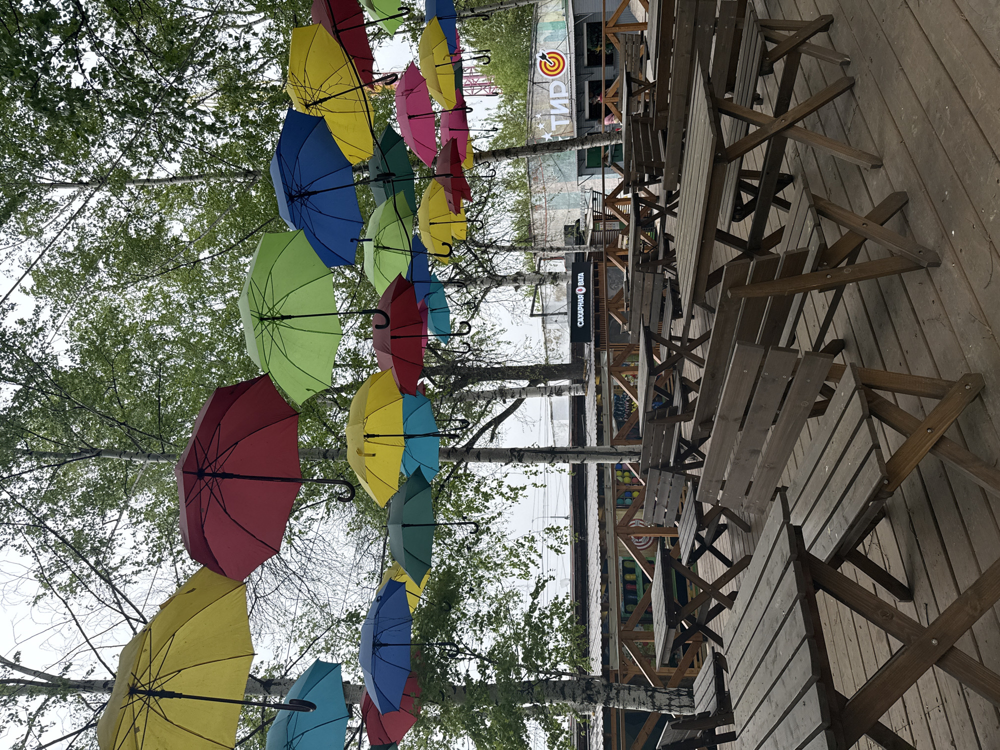 Outdoor scene in Izhevsk, Russia. Colorful umbrellas hang above wooden picnic tables and chairs on a wooden deck. Bare trees intermingle with the umbrellas. Buildings are visible in the background, including one with a sign in Russian.