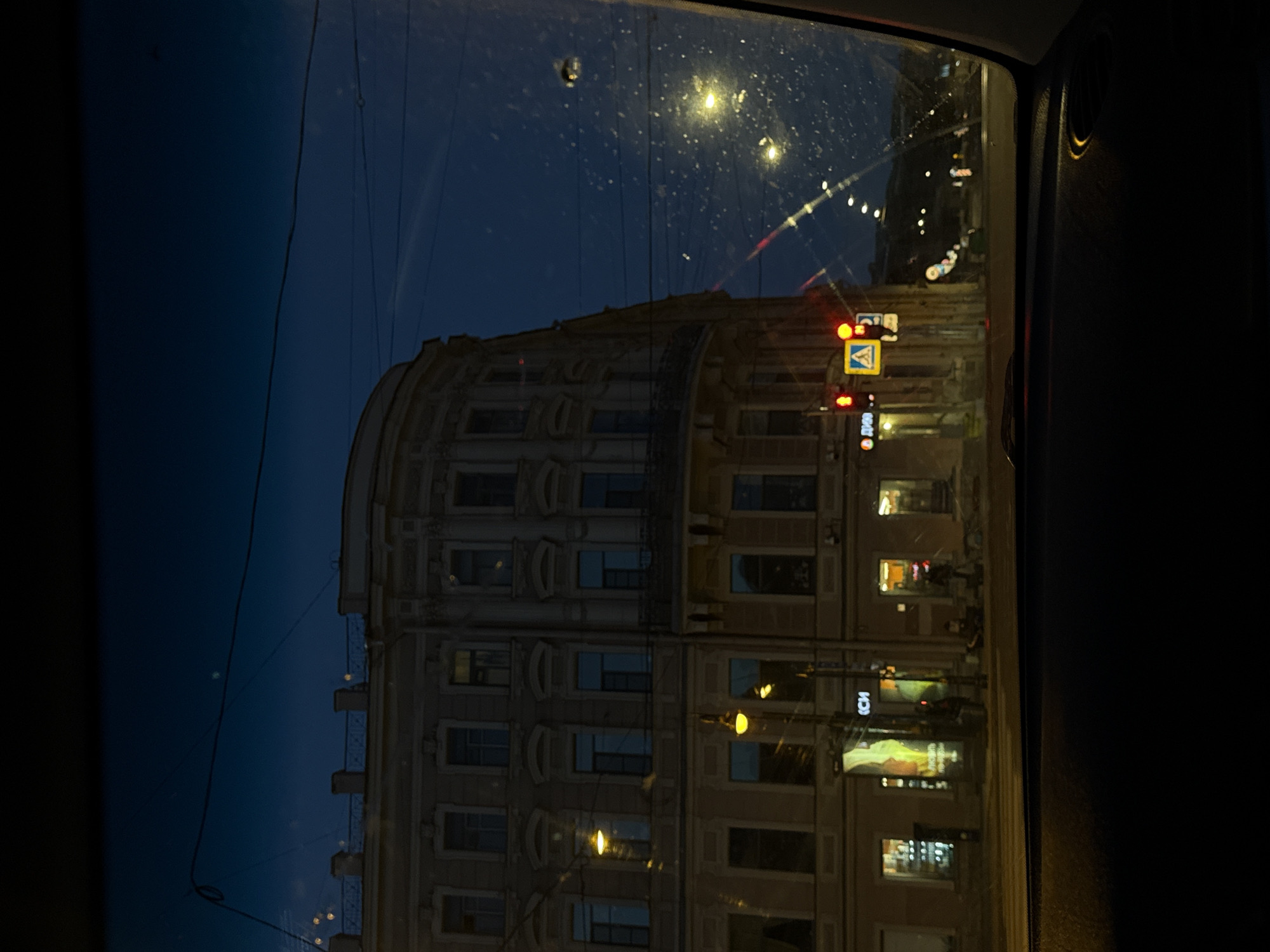 The image depicts a street view from inside a vehicle at dusk or night in Saint Petersburg, Russia. The facade of a large, ornate, historic building with multiple windows and architectural details dominates the left side of the frame. Streetlights cast a warm glow on the building and the street. A traffic light shows a red signal, and a pedestrian crossing sign is visible. In the background, the city lights and other buildings are dimly lit against the dark sky, with power lines crisscrossing overhead. The overall scene conveys a sense of urban nighttime ambiance.