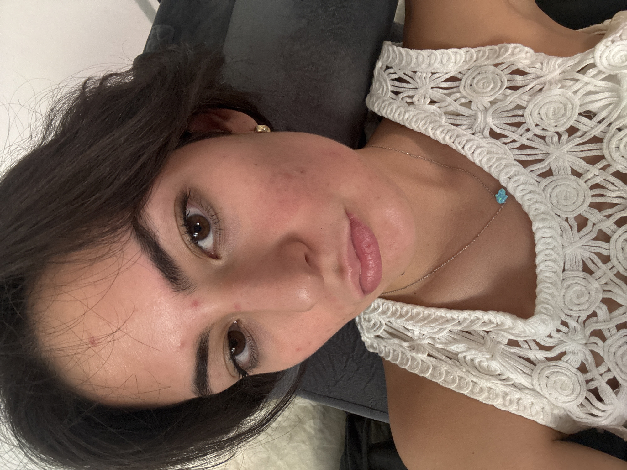 A woman with dark brown hair, brown eyes, and fair skin is looking at the camera. She is wearing a white crocheted top and a delicate silver necklace with a small, light blue charm. Her expression is neutral. She appears to be lying down on a dark grey surface. The location in the city of Puebla, Mexico is not discernible from this image.