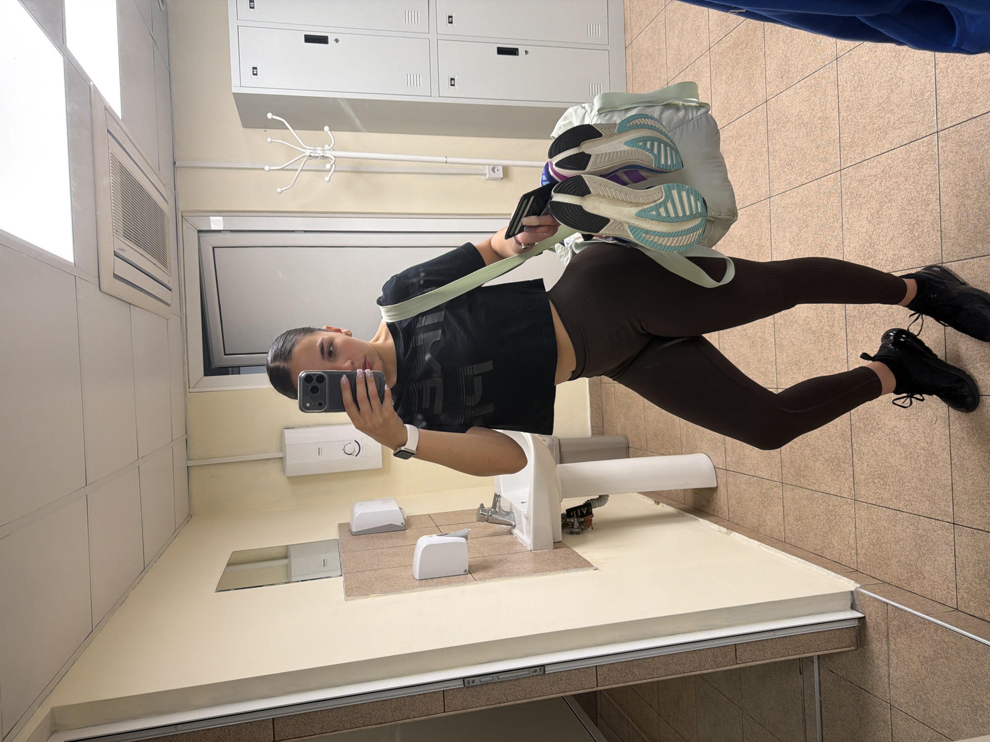 A person is taking a selfie in a restroom. The person is holding a mobile phone with their left hand and standing in front of a mirror. The person is wearing black athletic shoes, brown leggings, and a black athletic shirt. A gym bag containing athletic shoes is slung over their shoulder. The restroom has beige tiled floors and walls, a sink, a mirror, and a white door. In the background are a row of white lockers. The city of Gabrovo, Bulgaria is not identifiable from the image.