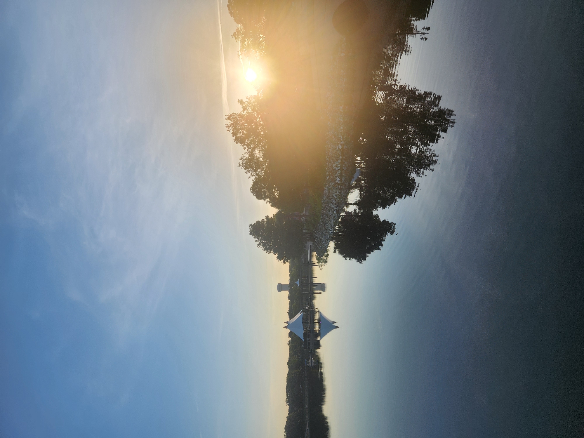 This is a vertical, wide-angle photograph of a lake at sunrise, taken from a slight elevation. The sky is clear and blue with wispy clouds. The sun is rising from the right, casting a warm golden glow that creates lens flare. The lake's surface is calm and reflective, mirroring the sky, trees, and structures on the opposite bank.
On the far side of the lake, there are trees lining the shore. In the middle ground, a white, tent-like structure with a pointed roof sits on a pier extending into the water, and its reflection is visible on the lake. Further back, a tall water tower is visible above the treeline. To the right of the image, there is a rocky shoreline and more trees, some of which are silhouetted against the bright sunrise. The overall impression is one of a serene morning scene.
This scene is likely at Cranes Roost Lake in Altamonte Springs, Florida, United States of America, given the presence of the distinctive tent-like structure and water tower, which are landmarks of the area.